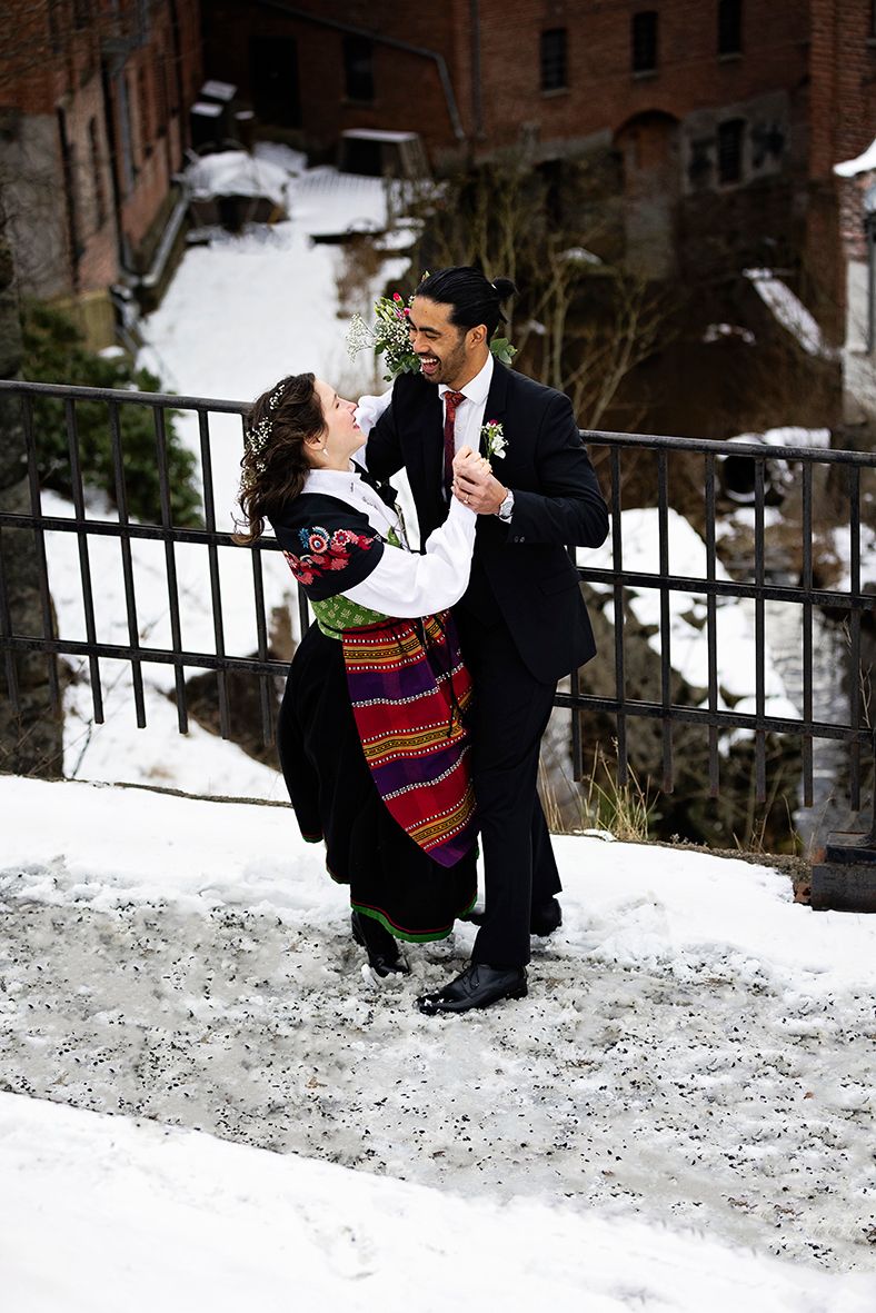 A couple playfully poses in the snow against a brick building backdrop.
