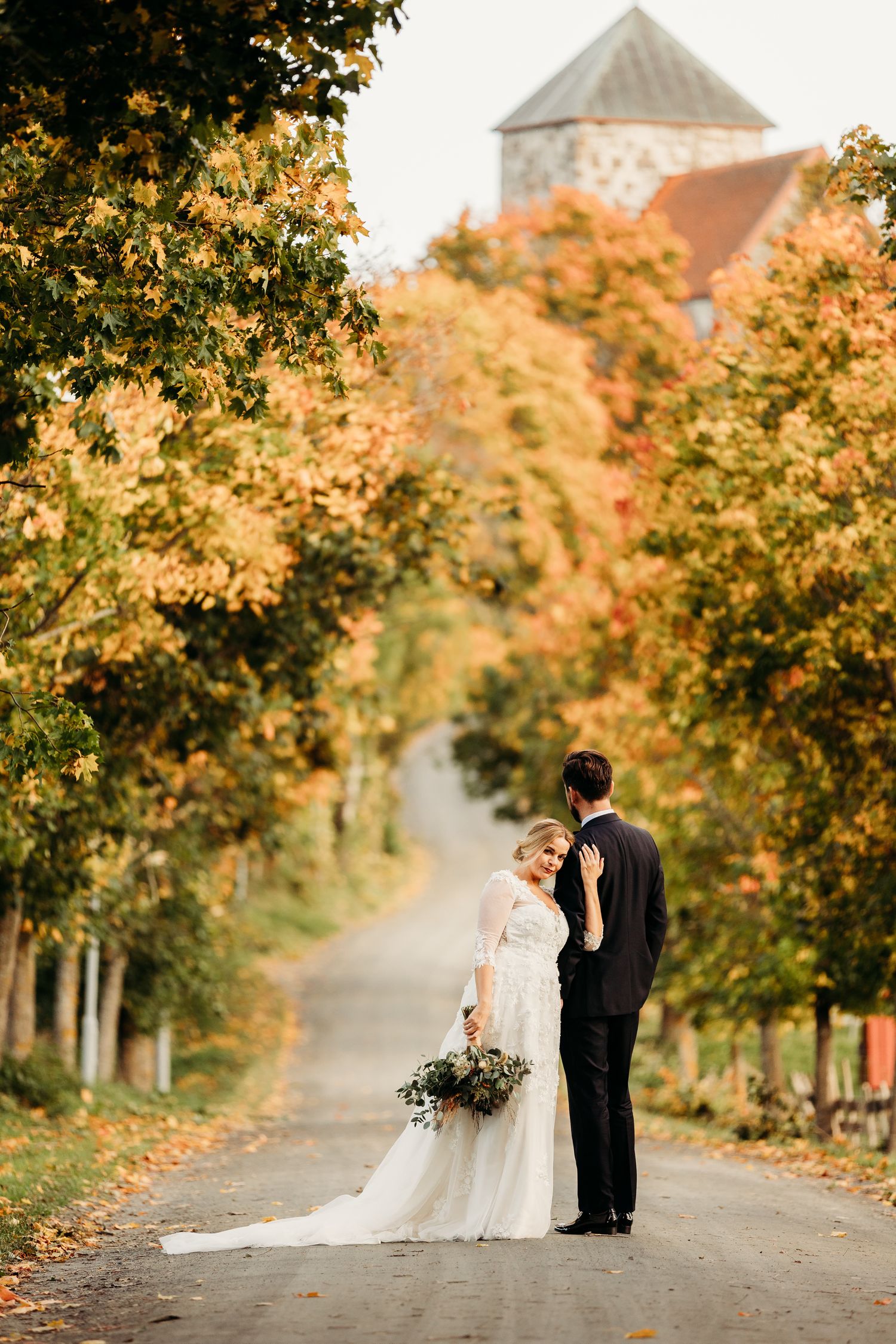 Wedding couple walks along tree-lined autumn path with golden foliage.