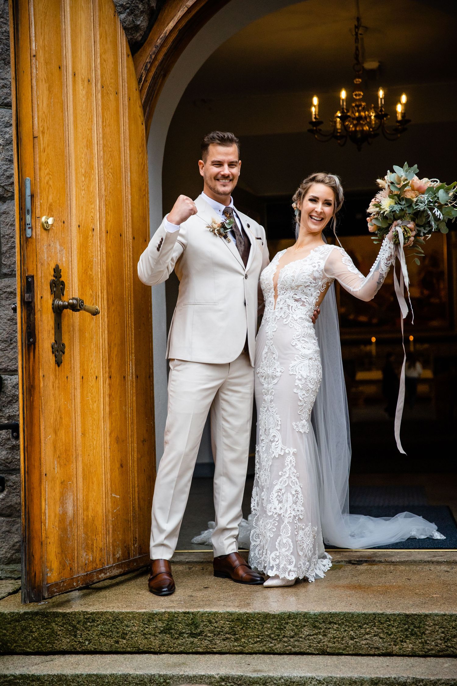 A wedding couple stands in front of large wooden church doors in white formal attire.