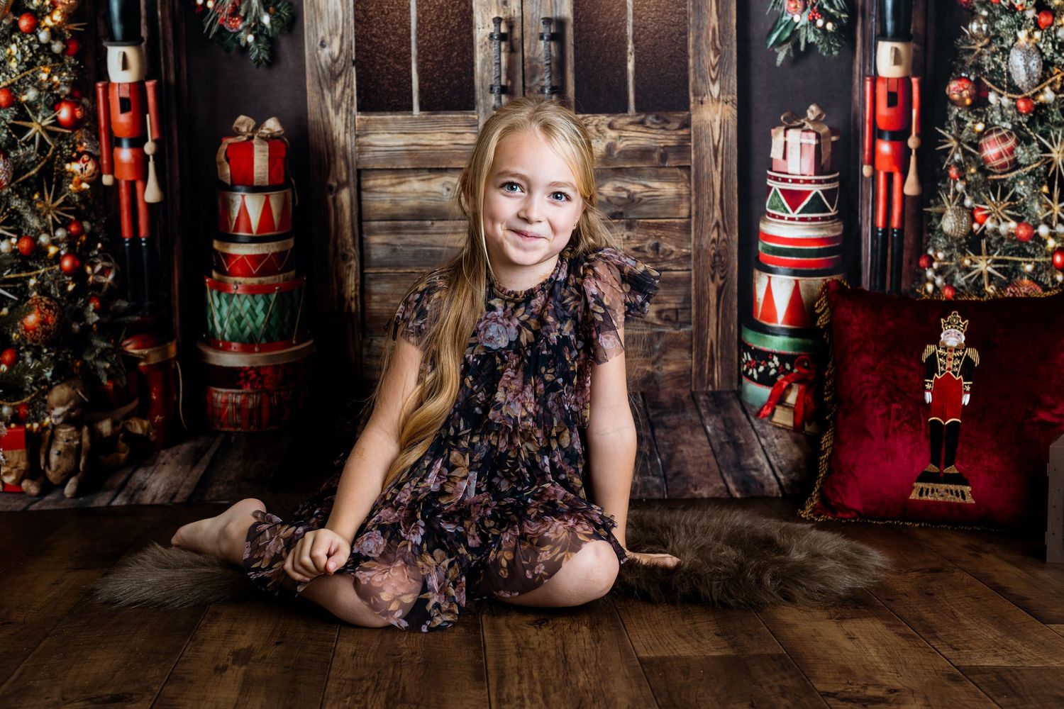 Holiday portrait with nutcracker decorations and rustic wooden backdrop featuring floral dress and festive setting.