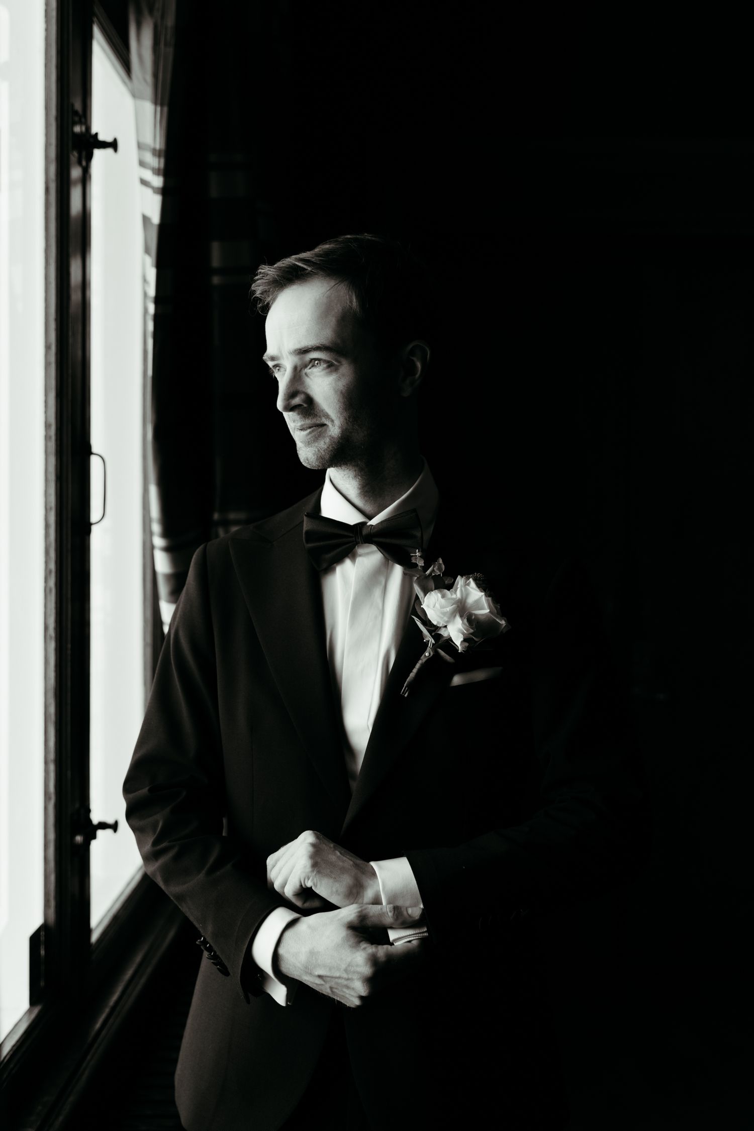 Dramatic black and white portrait of a groom adjusting his boutonniere in window light.