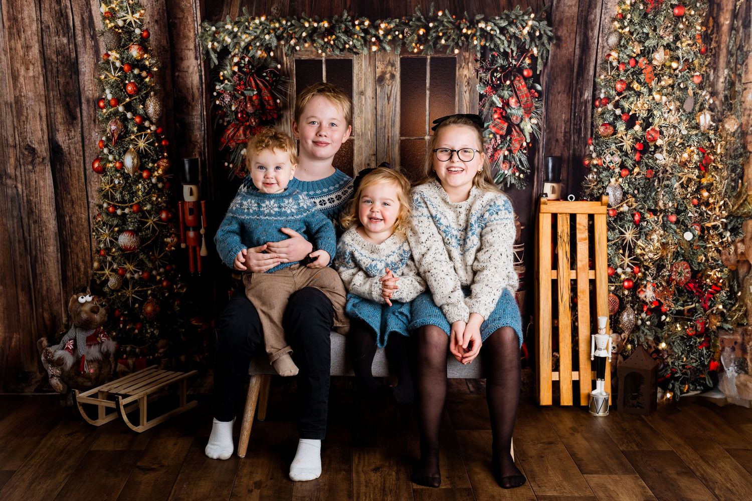 Fotograf Moss jul: Fire søsken i matchende strikkegensere sitter i en koselig og rustikk julebakgrunn med juletre og trevegg.