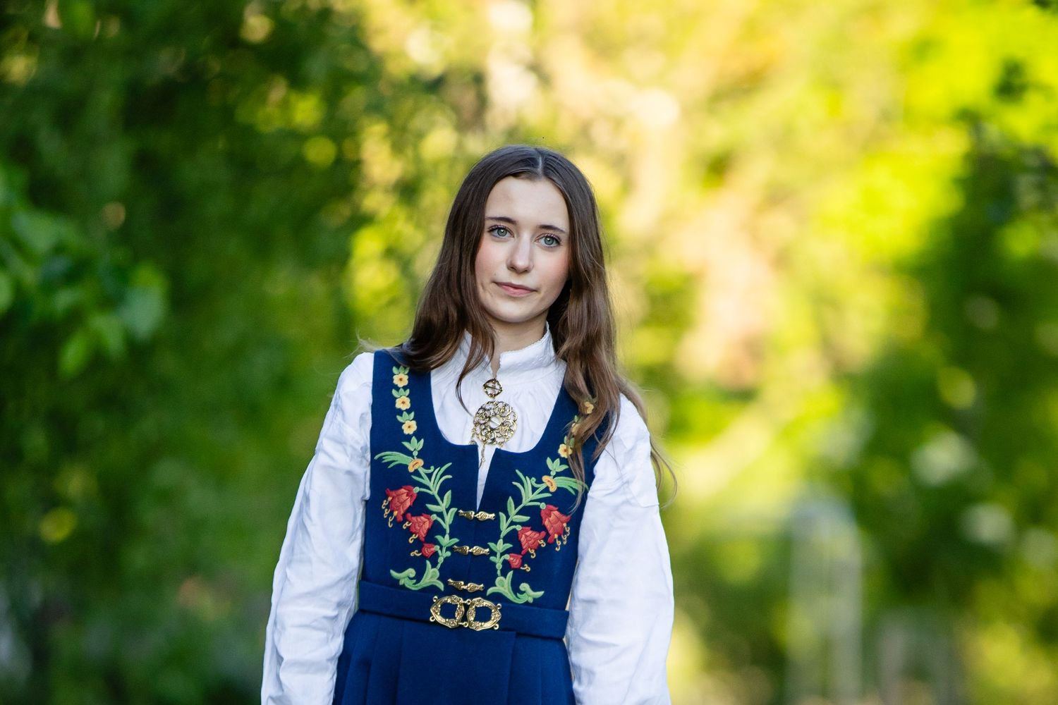 Traditional Norwegian folk dress with blue embroidered dress and white blouse shown in garden setting with flowers.