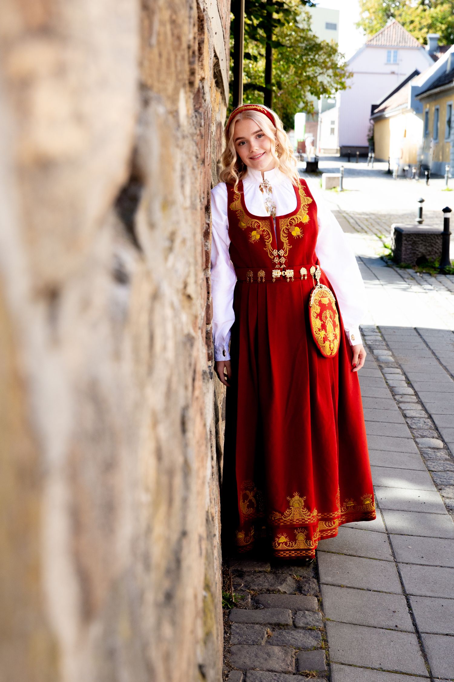 A full-length portrait in ornate red traditional dress with gold embroidery standing against stone wall.