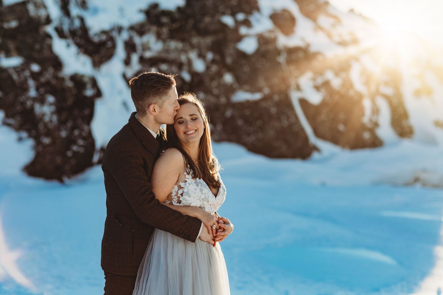 Couple sharing intimate moment during winter wedding photos with sunlit snow backdrop.