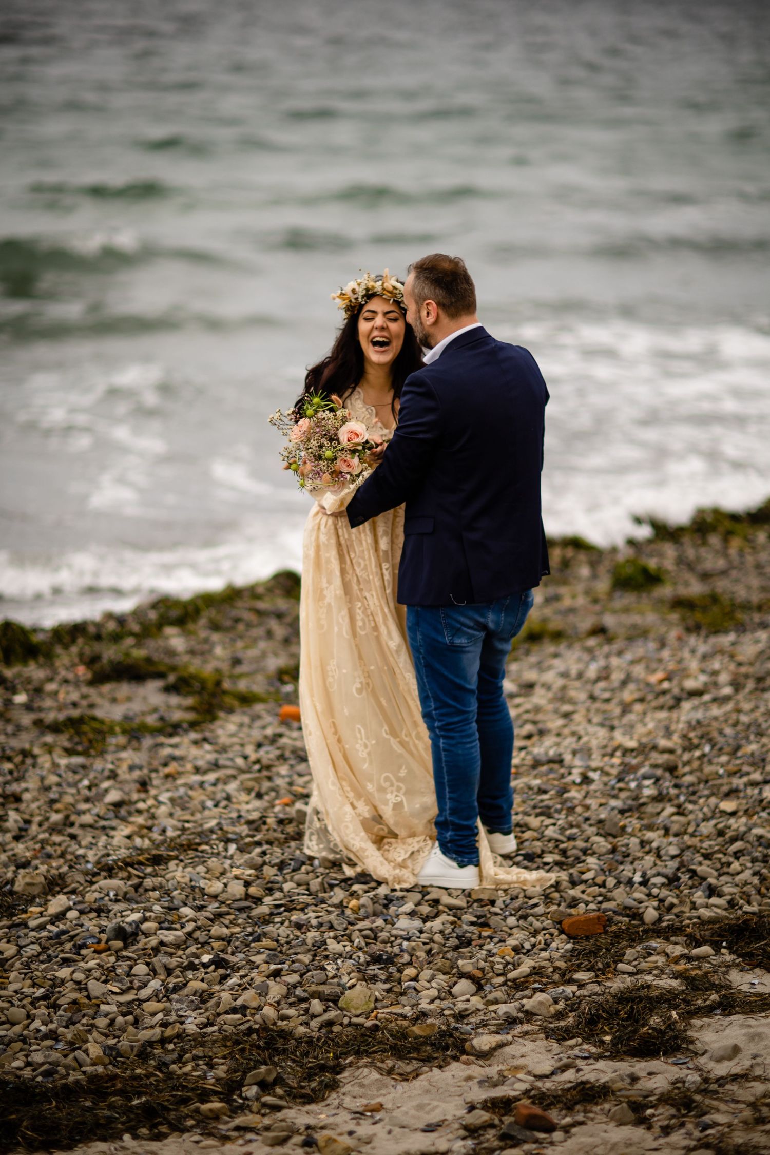 Romantic seaside wedding photos with couple embracing on pebble beach.