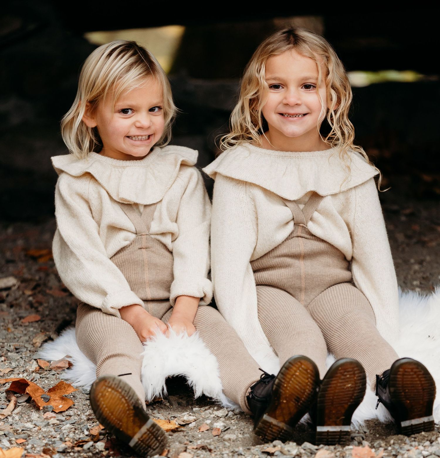 Siblings in coordinating beige outfits sitting together on ground with matching boots.