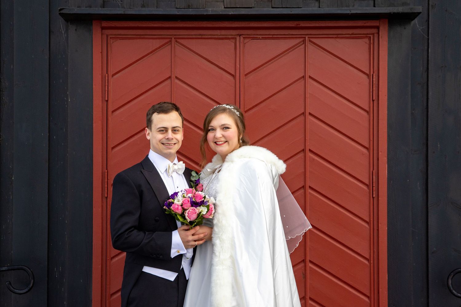 Wedding couple stands before red chevron door with bride in white cape and groom holding pink bouquet.