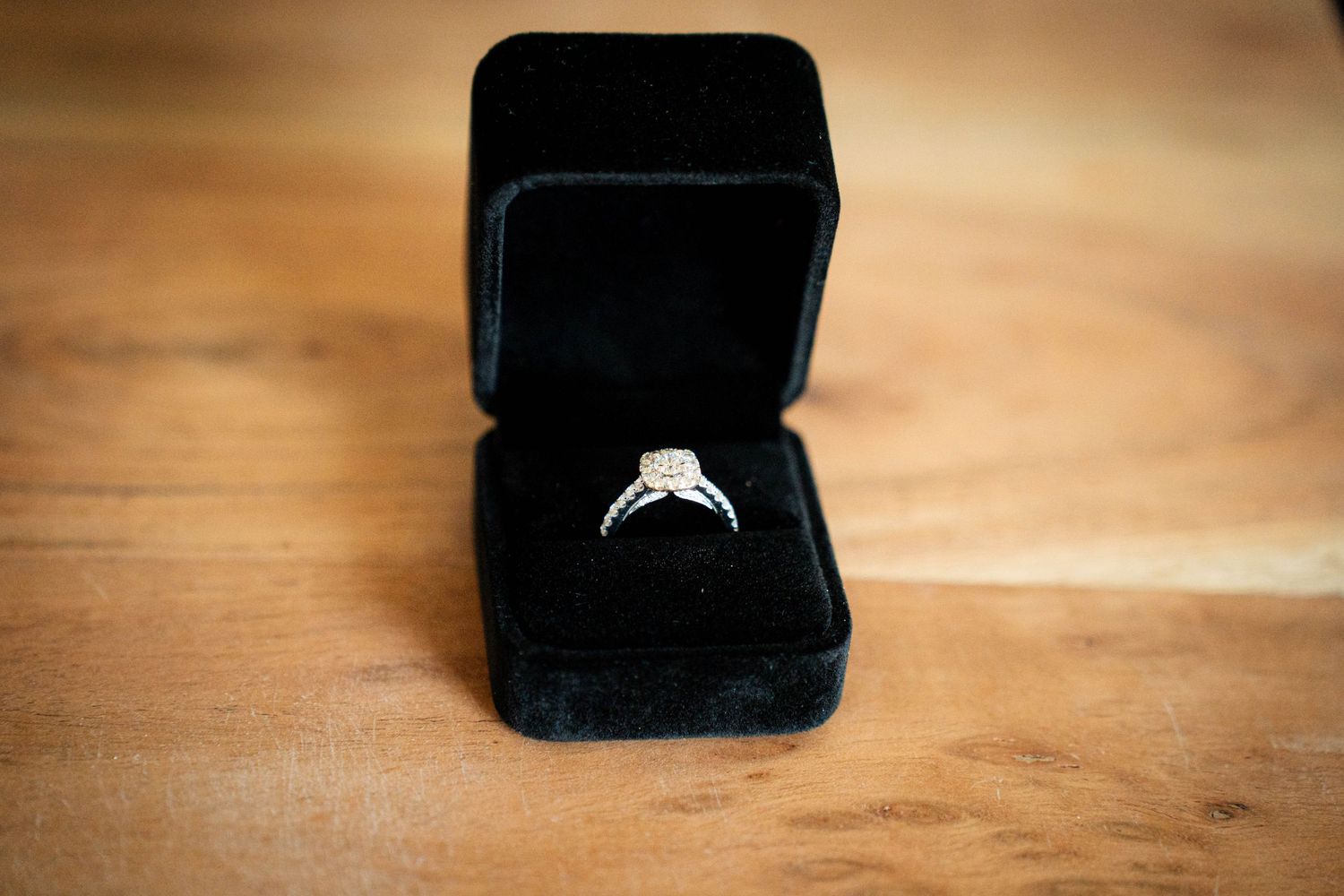 Diamond halo engagement ring displayed in open black velvet jewelry box on wooden surface.