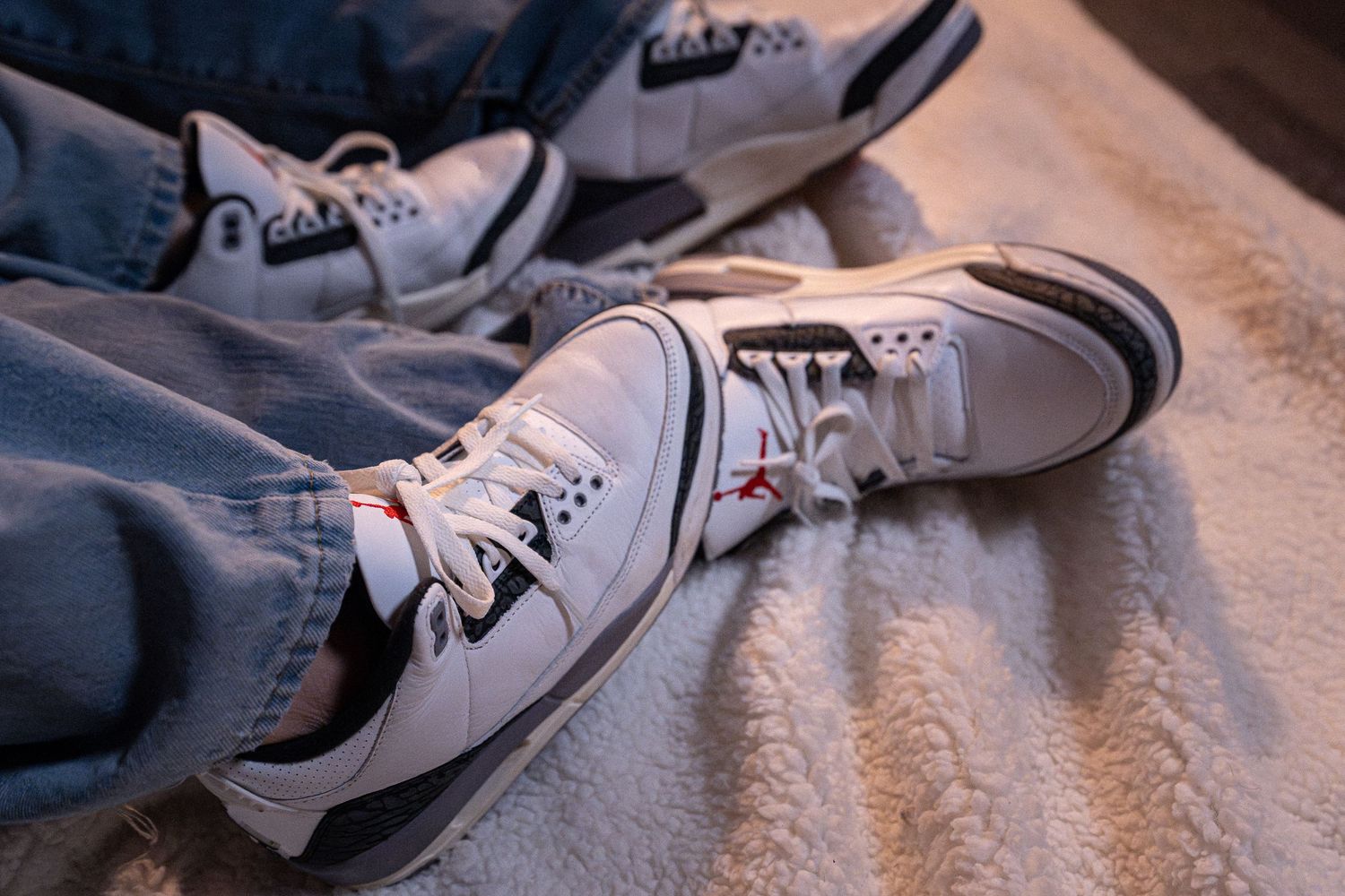 White Air Jordan 3 sneakers worn with light blue jeans on a soft textured surface.