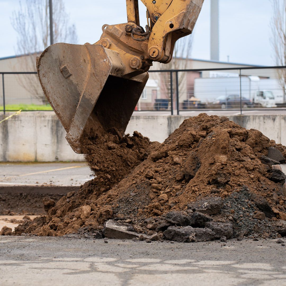 Yellow excavator bucket depositing a pile of brown dirt and broken asphalt on a construction site.