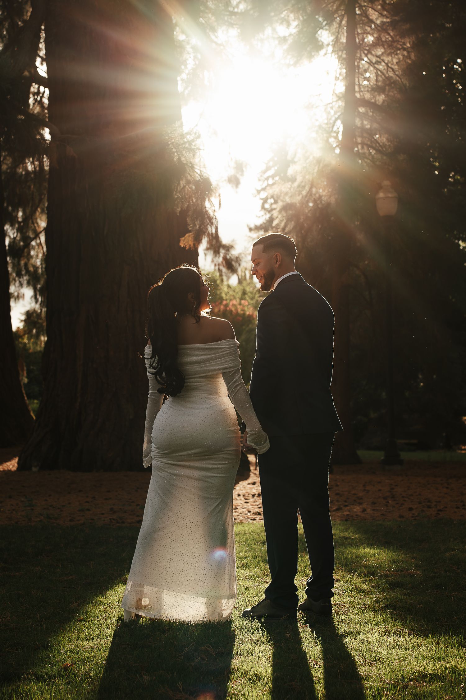 Bride and groom holding hands in a forest, backlit by golden sunlight streaming through tall trees.