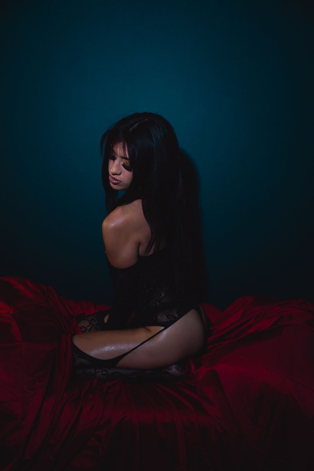 A woman in black lingerie sits on red satin sheets against a dark teal background.