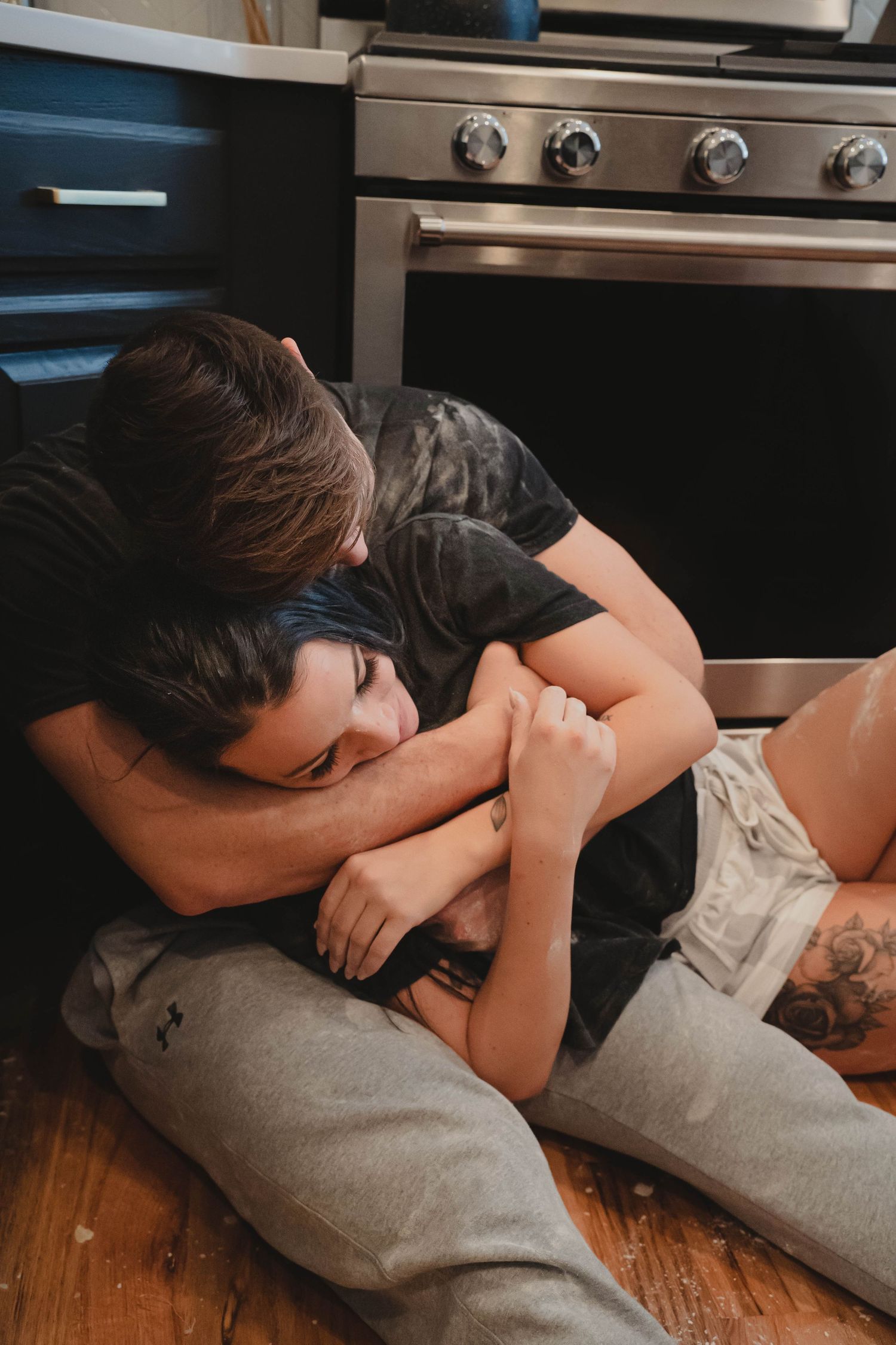 Couple sharing a tender embrace on kitchen floor with tattoos and casual clothing.
