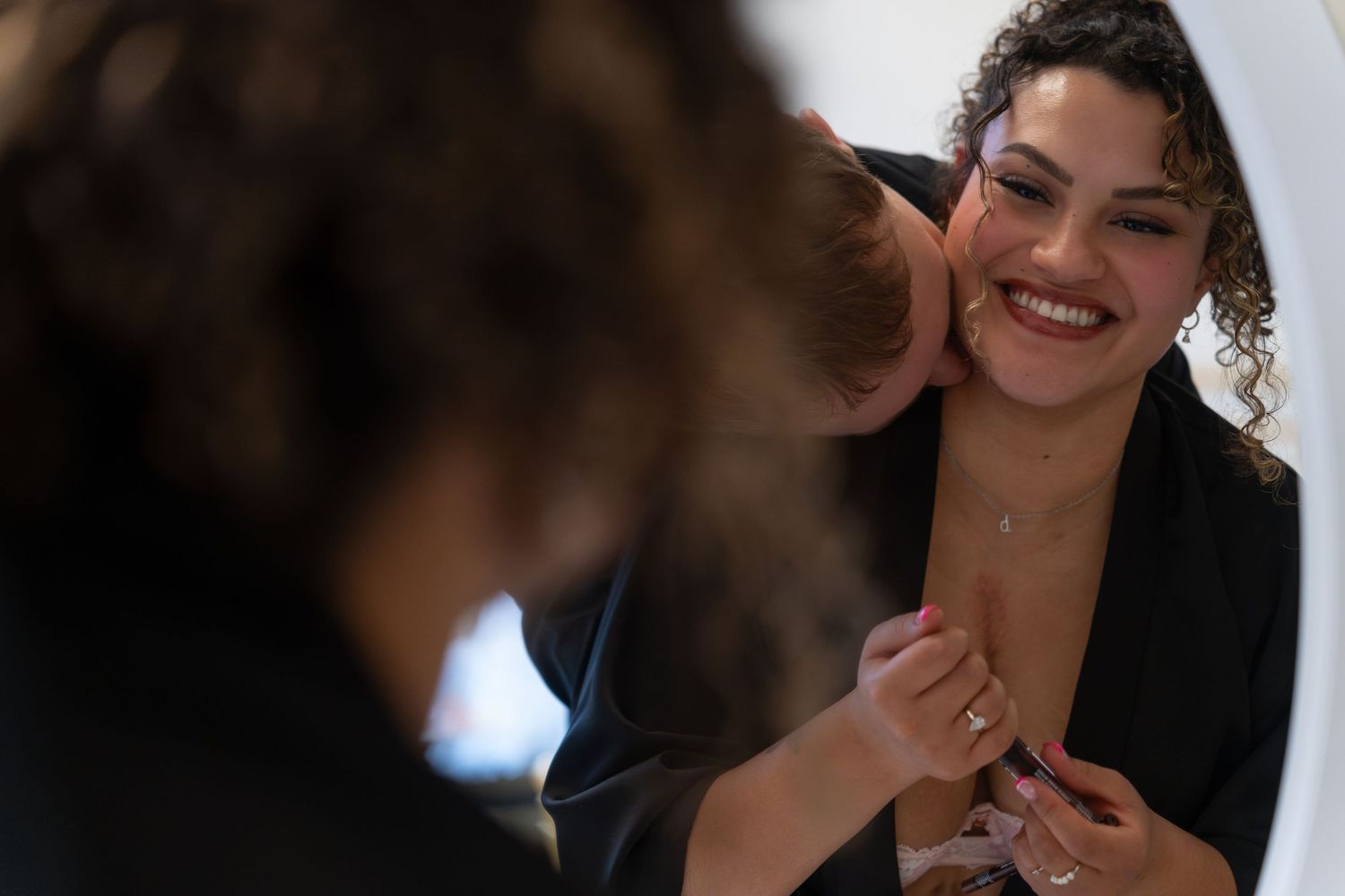 A smiling woman in a black robe looks in a mirror while someone adjusts her top.