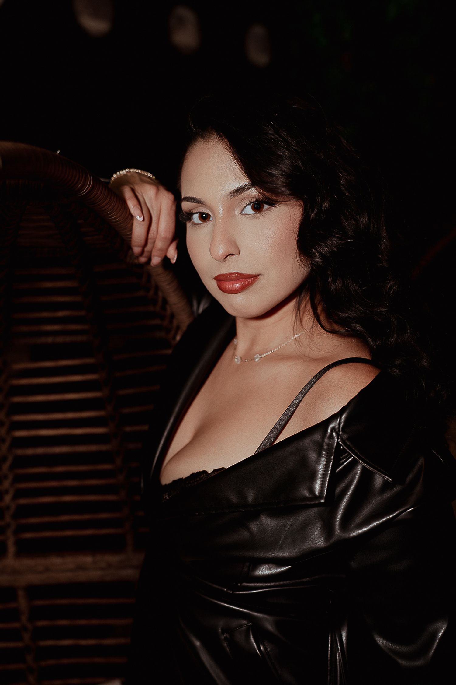 A brunette woman in a black leather jacket and low-cut top poses in a dimly lit outdoor setting.