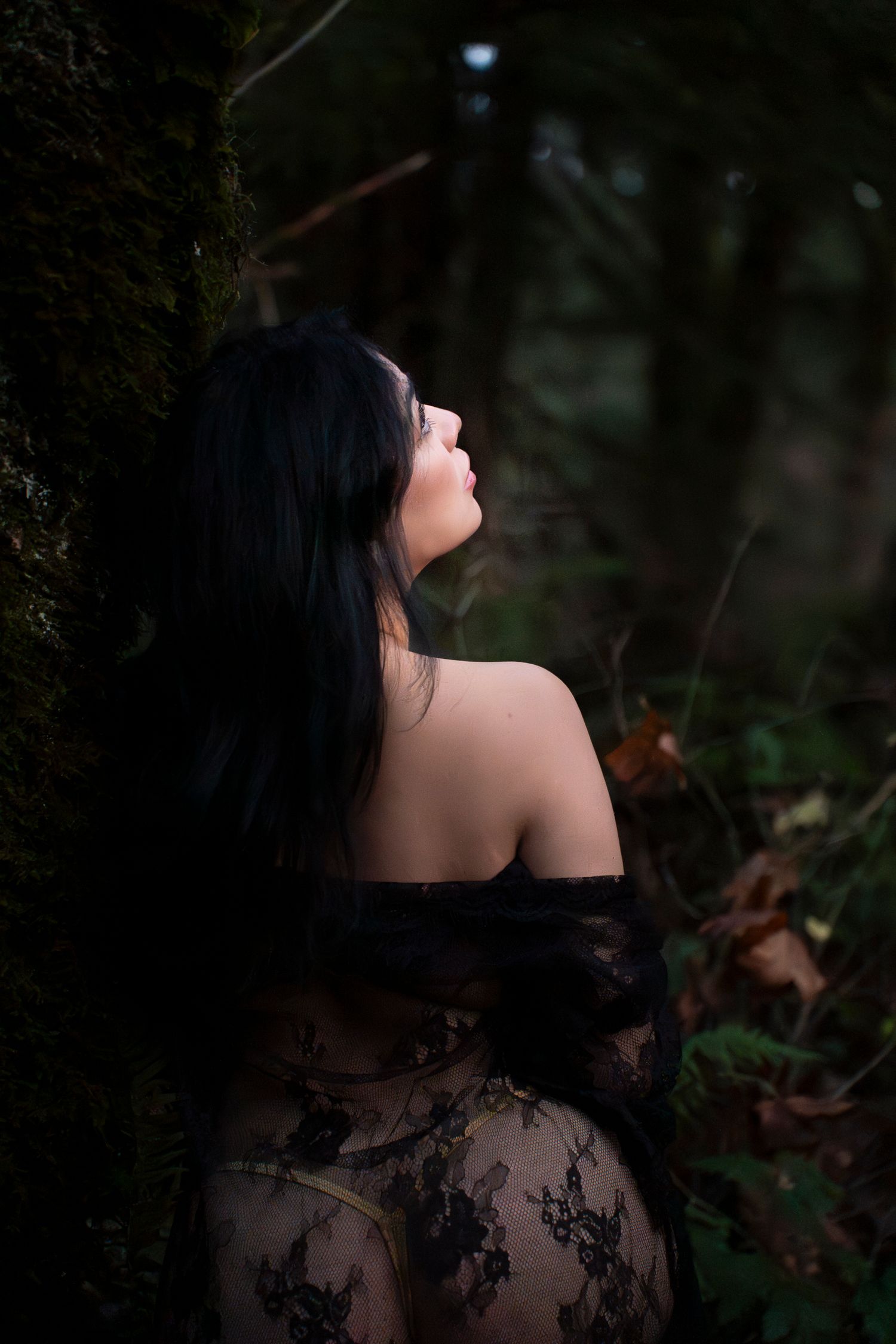 A woman in black lace clothing leans against a moss-covered tree in a dark forest setting.