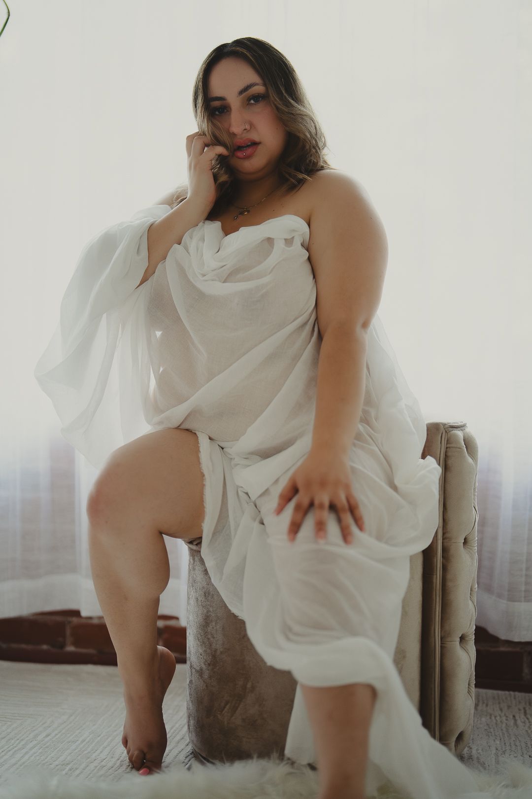Woman wrapped in white sheer fabric sitting on rustic chair in bright airy room.
