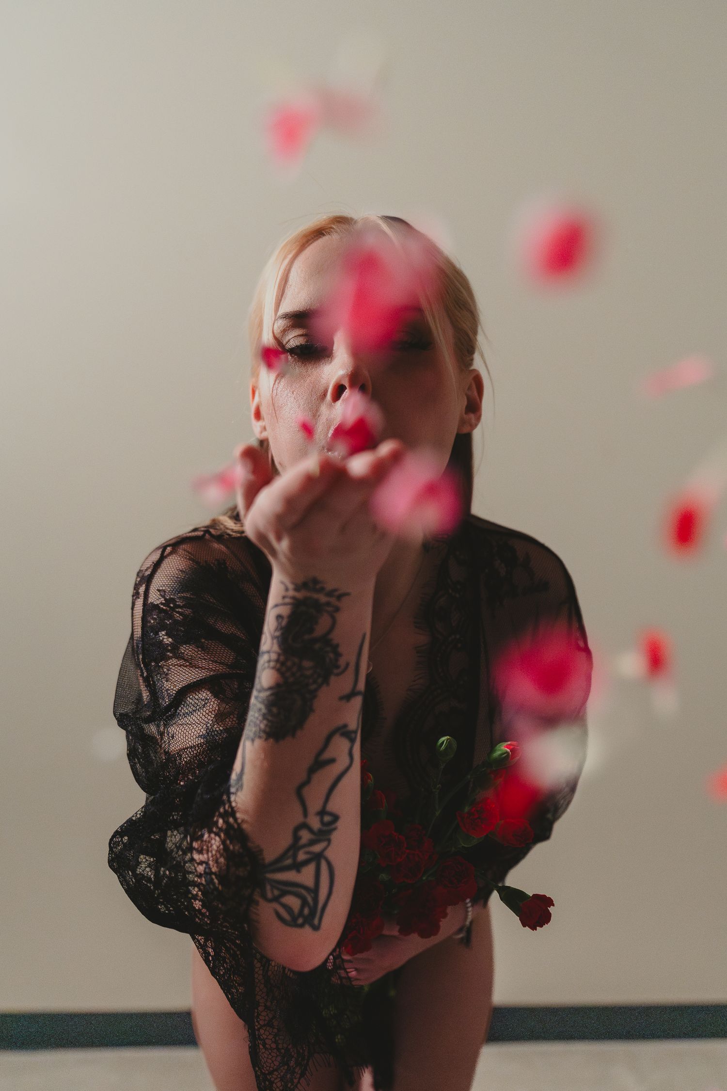 Blonde tattooed woman in black lace bodysuit blowing pink rose petals in a studio setting.
