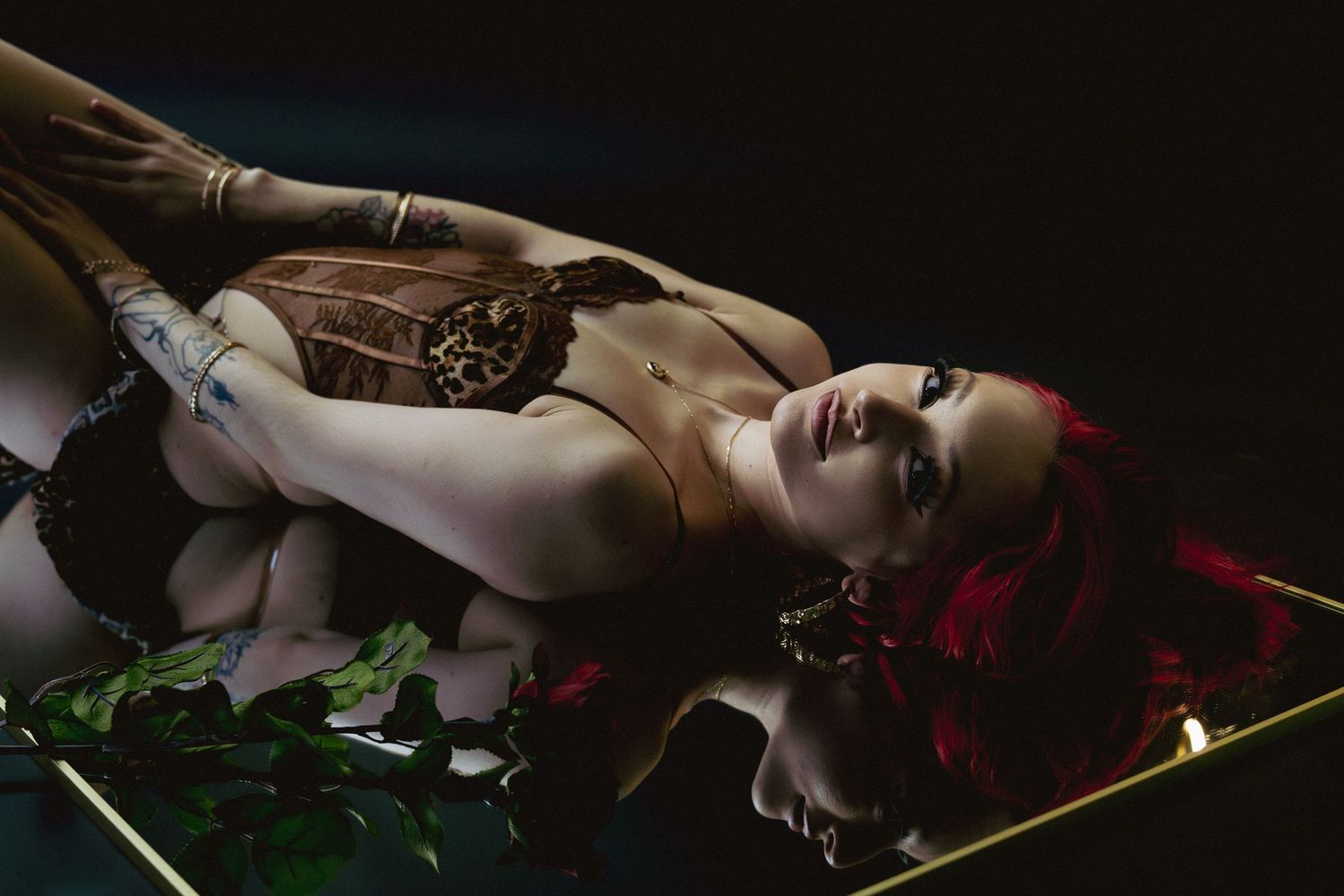 A woman with vibrant red hair lies on a reflective surface wearing a leopard print corset with green foliage.