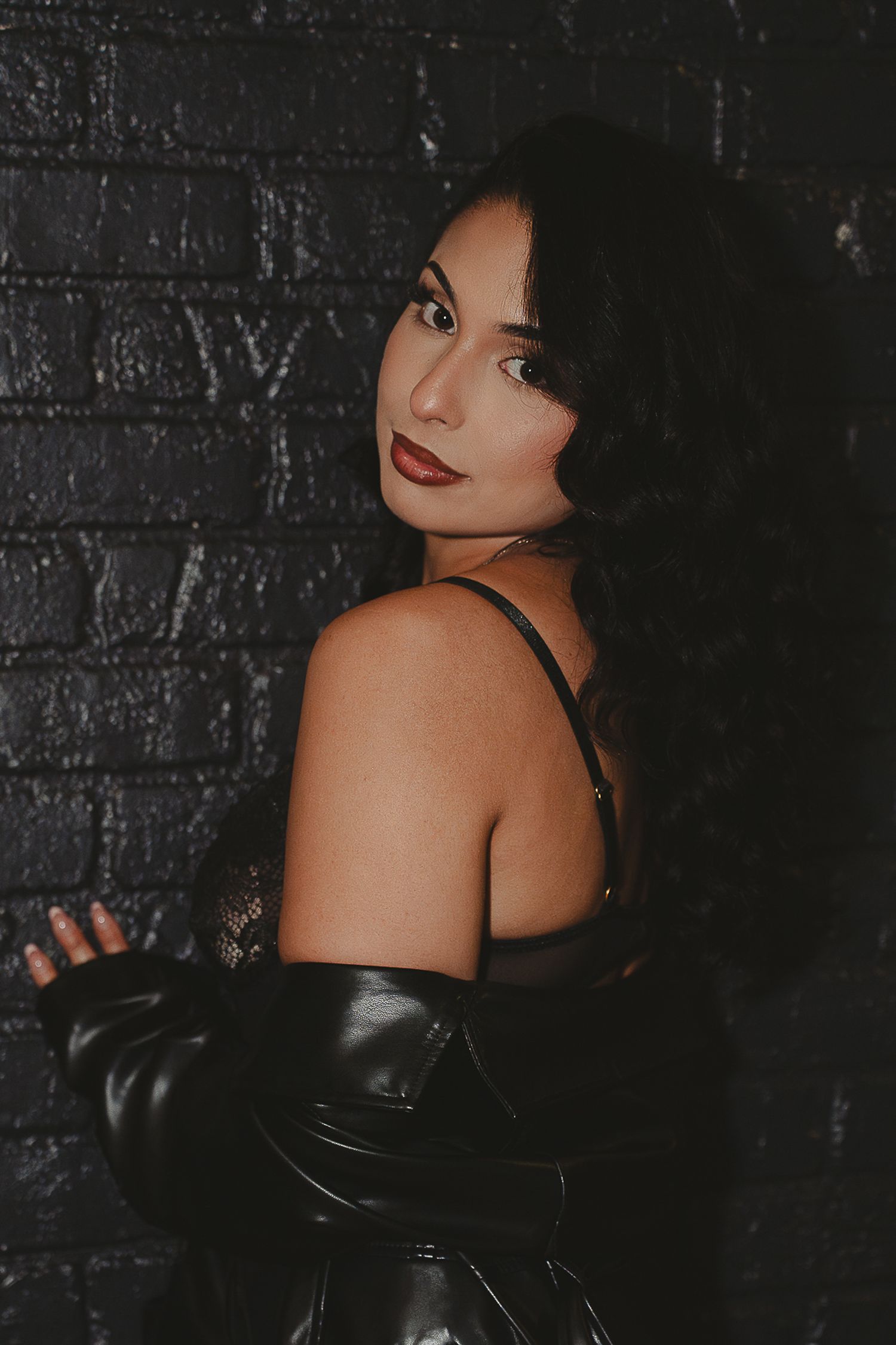 Dark-haired woman in black leather outfit posing against a dark brick wall with red lips.
