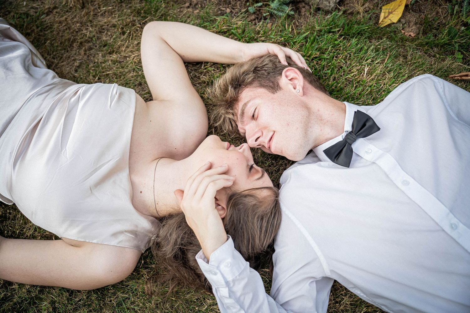 A young couple lies on grass, gazing at each other in wedding attire.