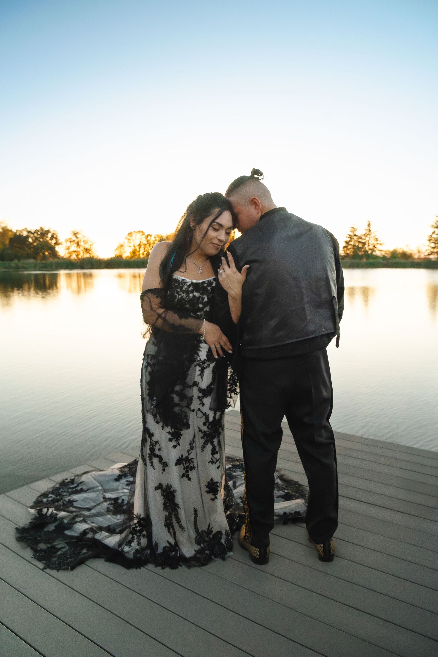 A bride in a black lace gown and groom in a leather jacket embrace on a dock at sunset.