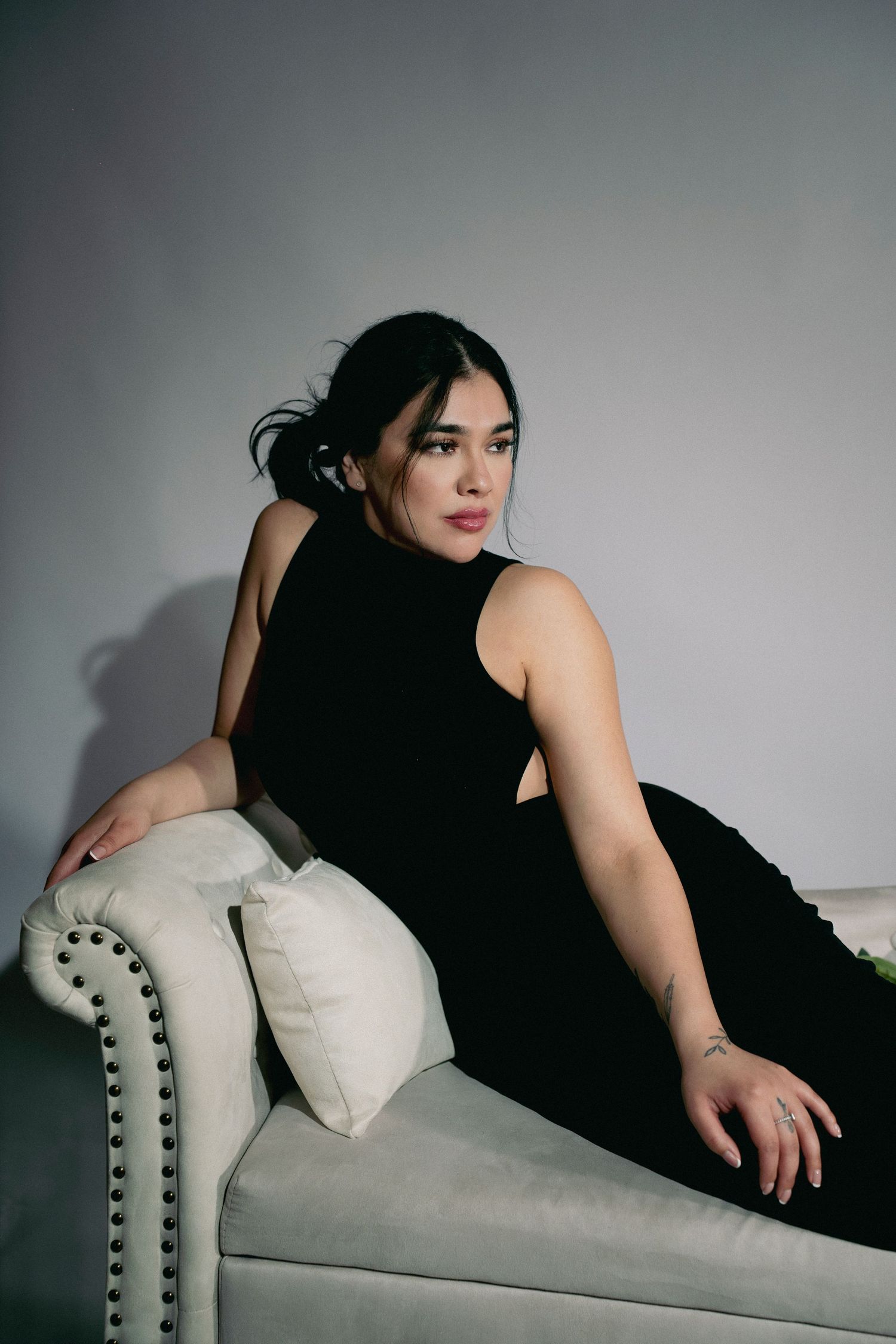 A dark-haired woman in a black cutout dress poses gracefully on a white velvet chaise lounge in a studio.