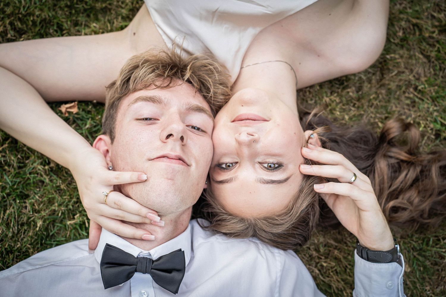 A young couple lies head-to-head on grass, smiling, with the man wearing a black bow tie.