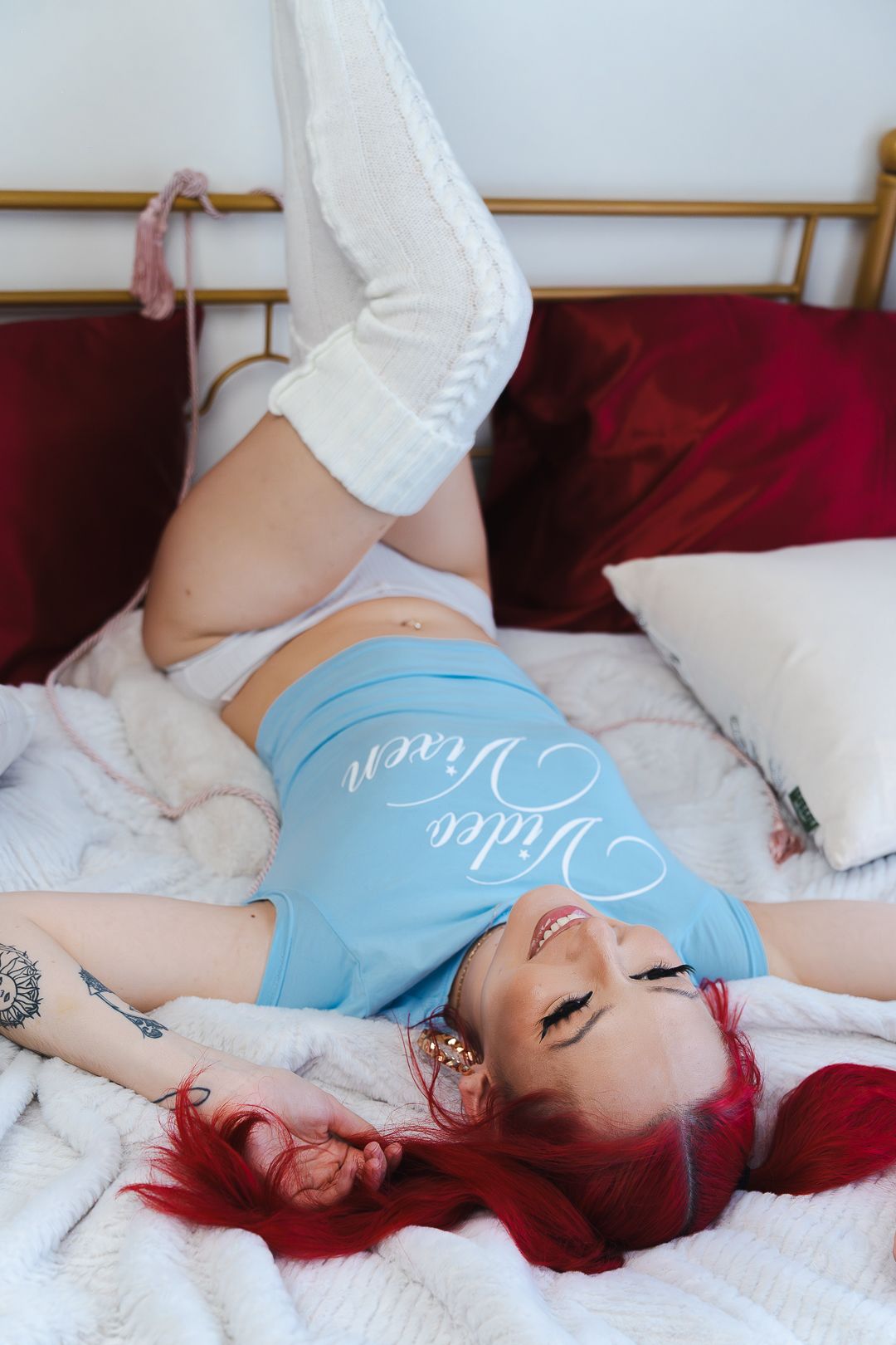 Red-haired woman in blue crop top and white pants lying on bed smiling.
