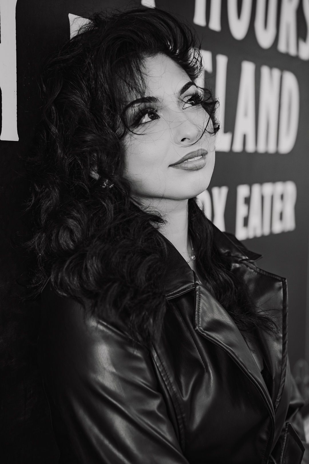 A woman with voluminous curly dark hair wearing a leather jacket poses in black and white photo.