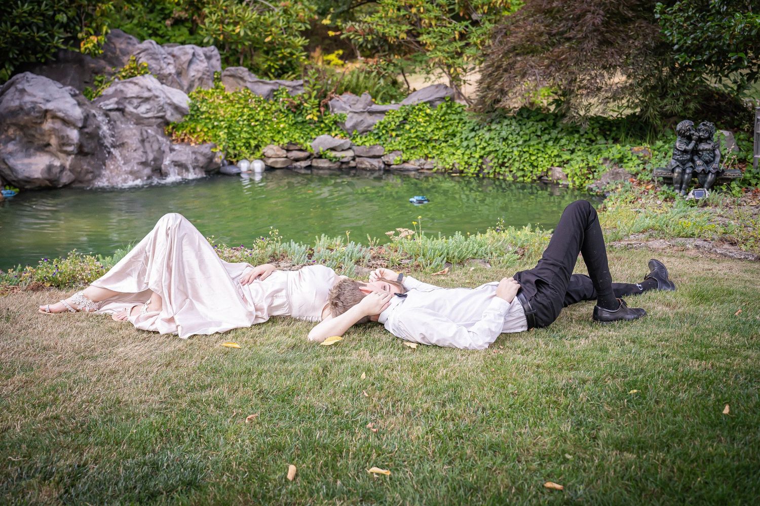A bride and groom lay playfully on green grass near a serene garden pond on their wedding day.