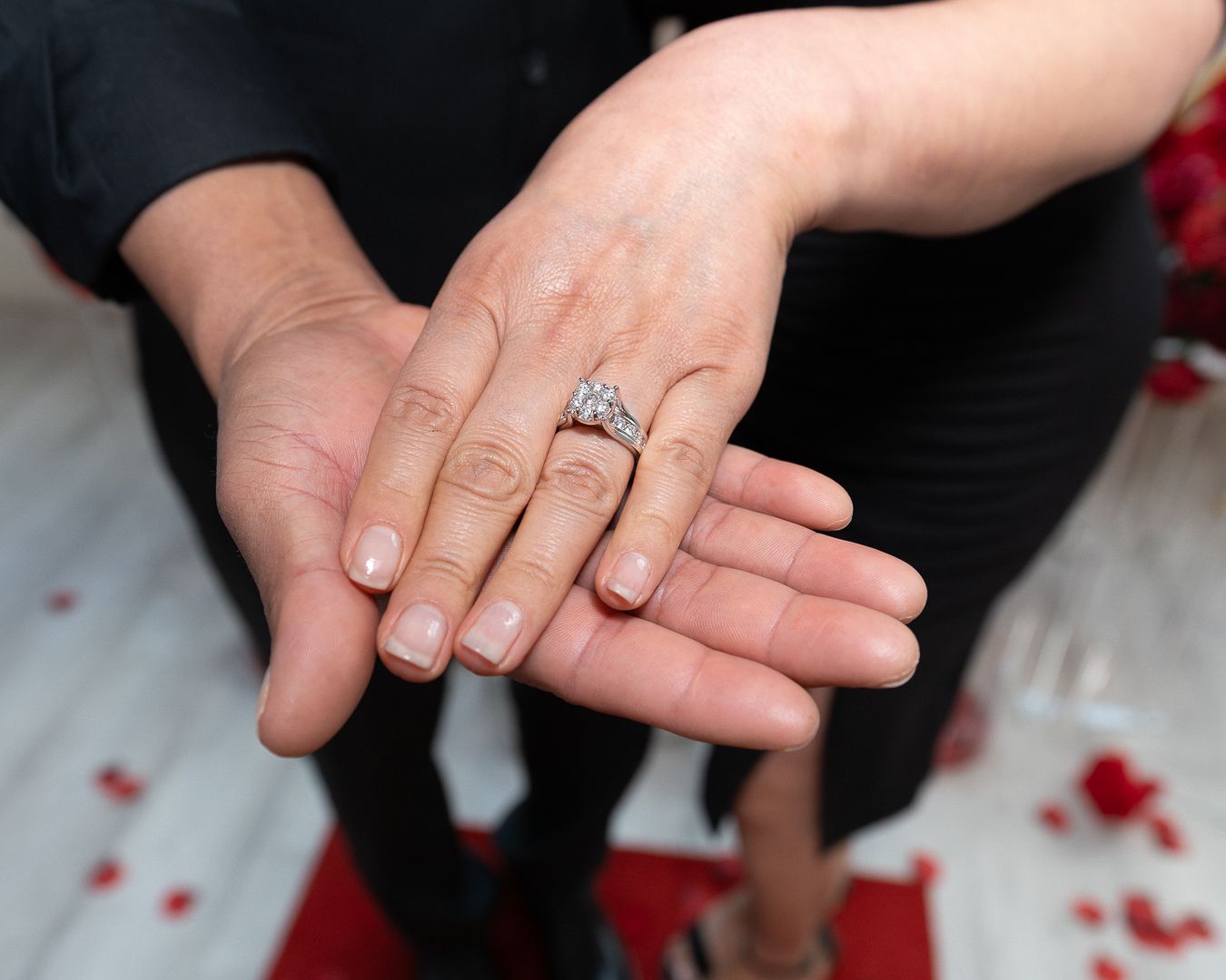 Couple displaying engagement ring on joined hands with red rose petals scattered on white floor.
