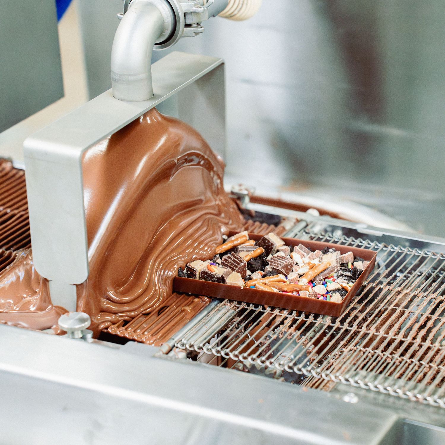 Liquid milk chocolate pouring over nuts and candy pieces on a factory conveyor belt.