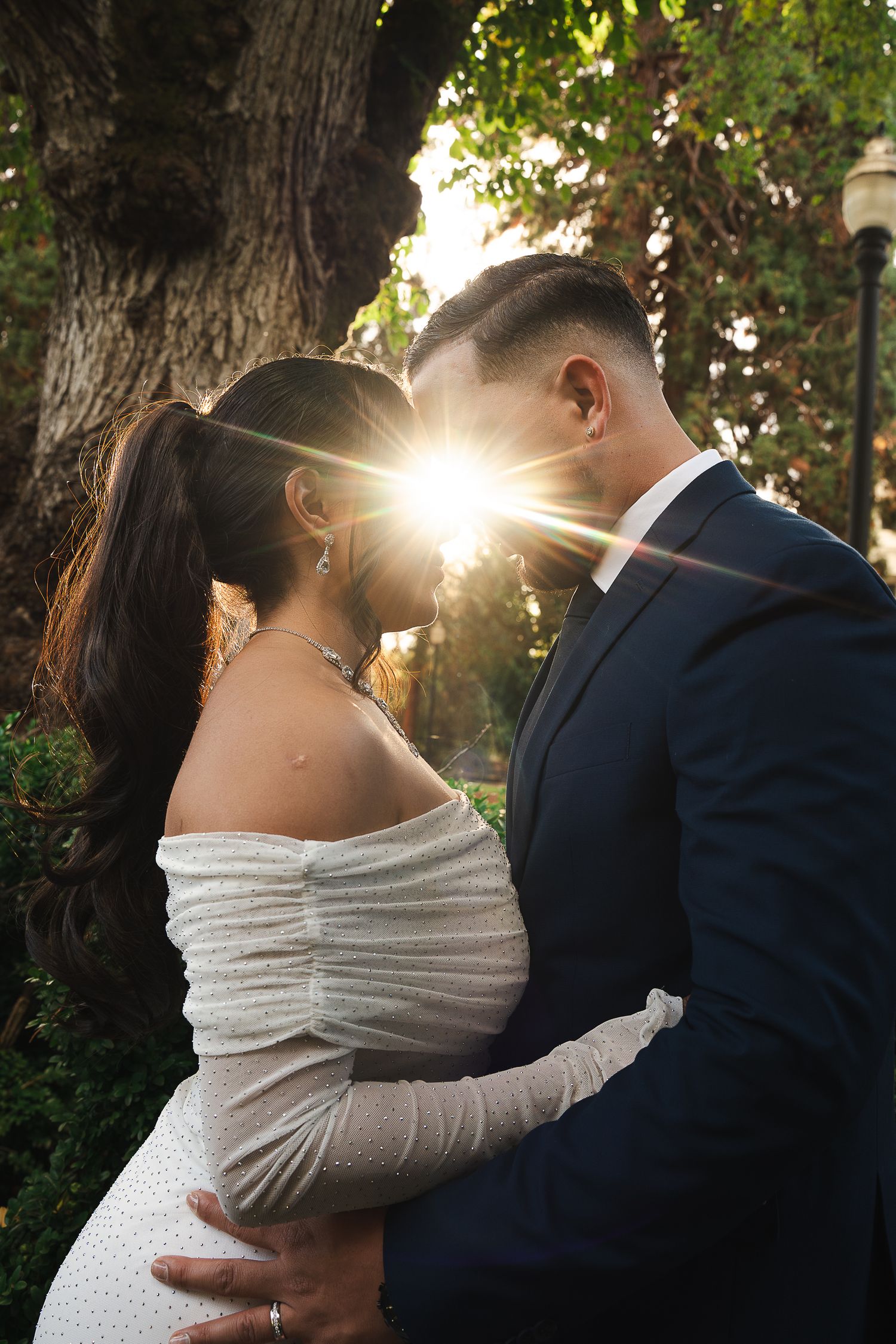Couple sharing a romantic kiss backlit by golden sunset rays in a lush garden, bride in off-shoulder white gown in Beaverton, Oregon.