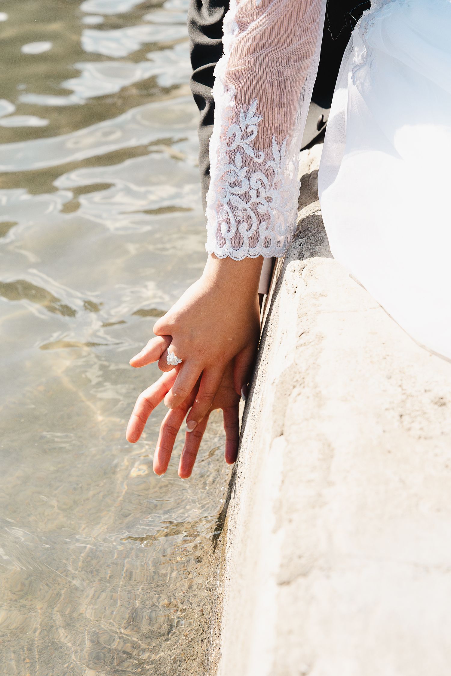 Bride in lace wedding dress touching water with ringed hand on a sunlit stone ledge at the Portland Peninsula Park Rose Garden.