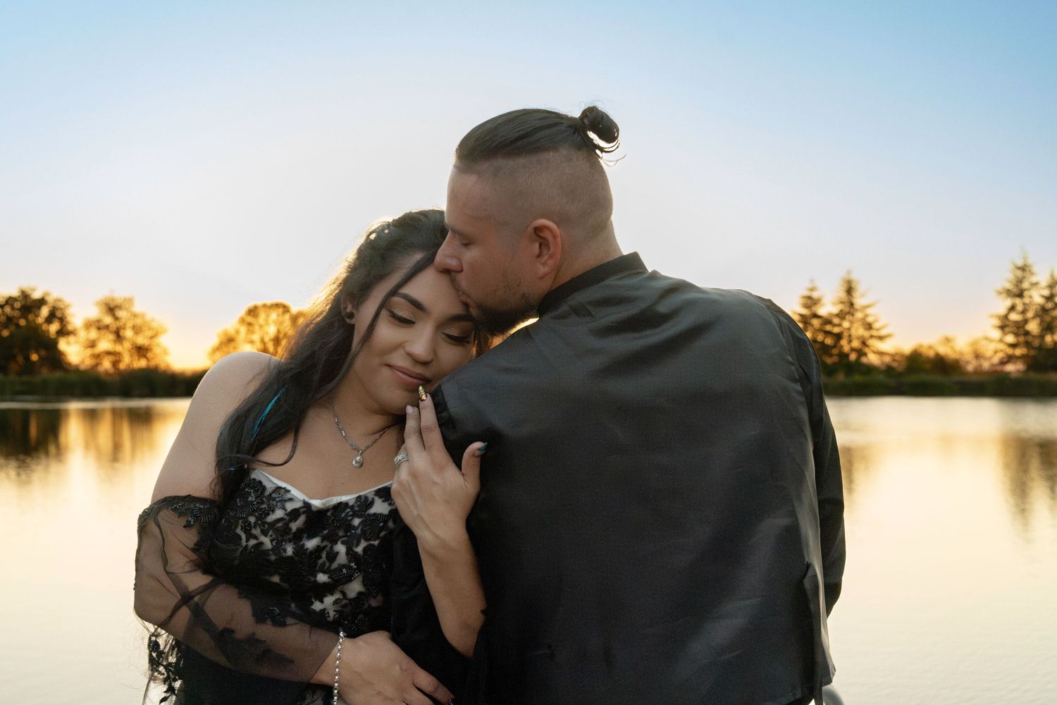 A couple embraces tenderly by a serene lake at golden hour sunset.