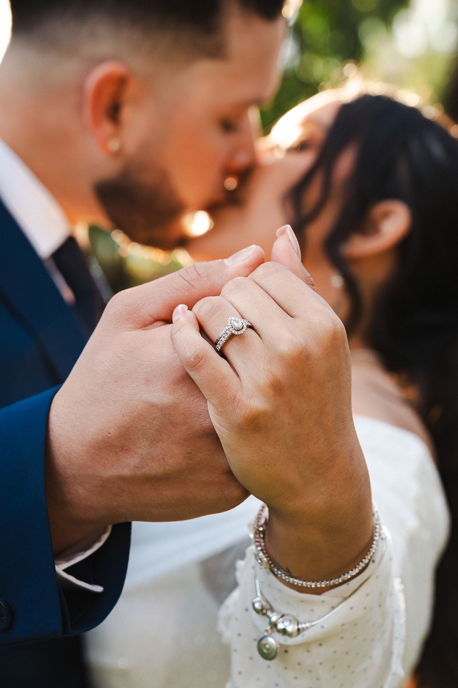 Close-up of bride's hand showing diamond engagement ring and wedding band while couple kisses softly.