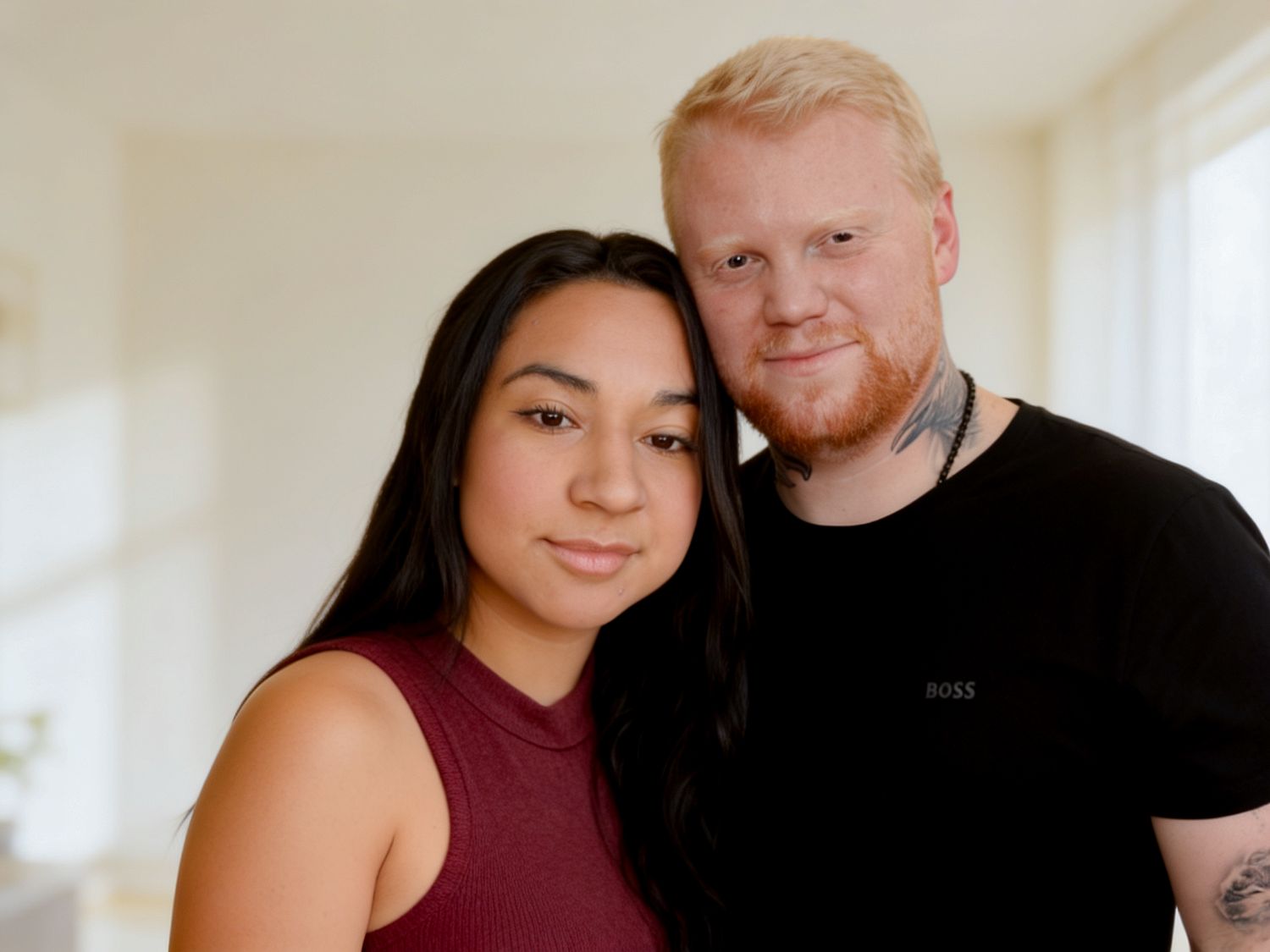 A couple posing closely together in a bright room, the man in a black Boss shirt and the woman in a burgundy top.