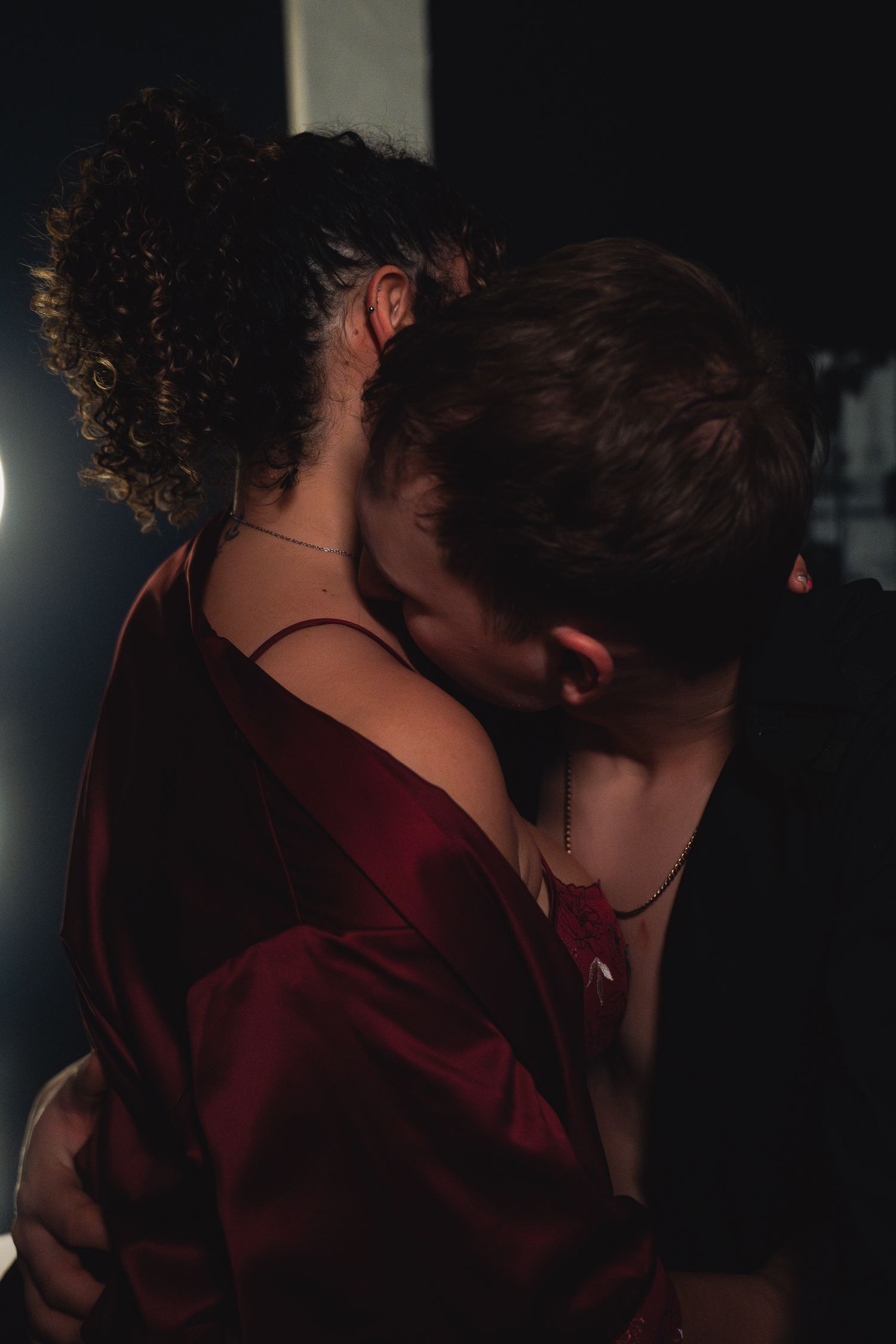A couple shares a romantic embrace in dim lighting, with the woman wearing a deep red satin dress.