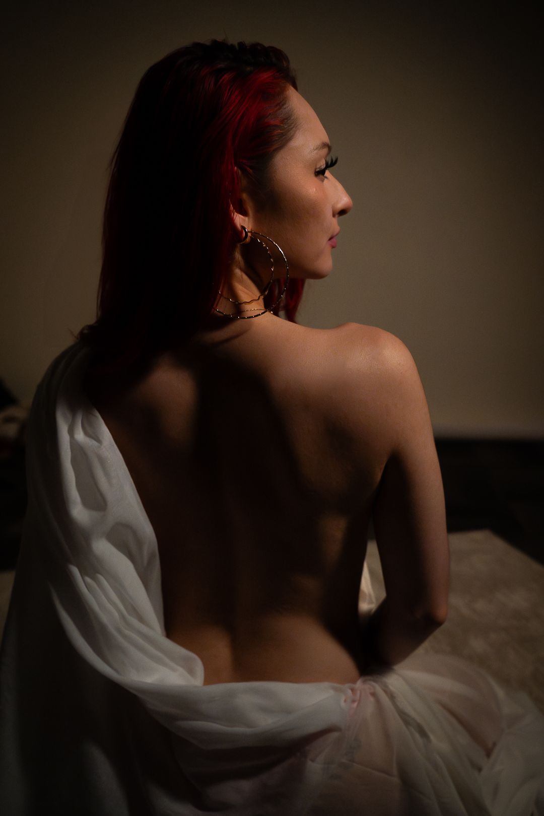 Woman with red hair sitting on a bed draped in white fabric showing bare back in dramatic lighting.