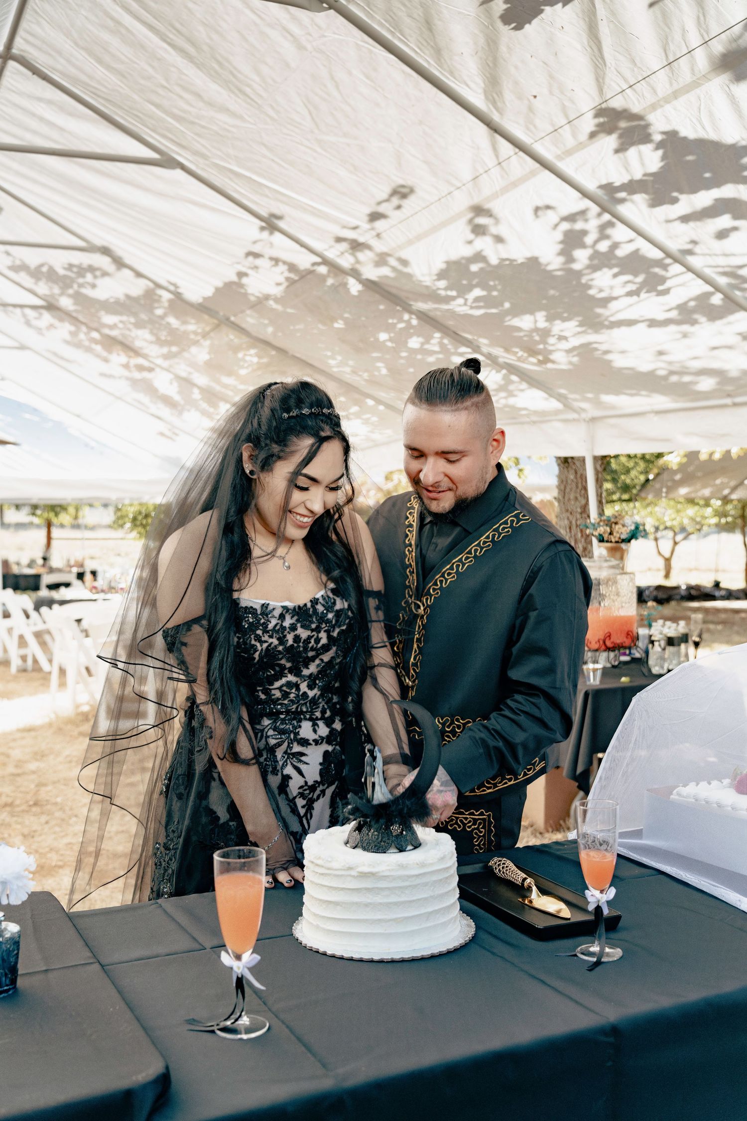 Gothic wedding couple in black attire cutting a white cake decorated with dark flowers under a tent.