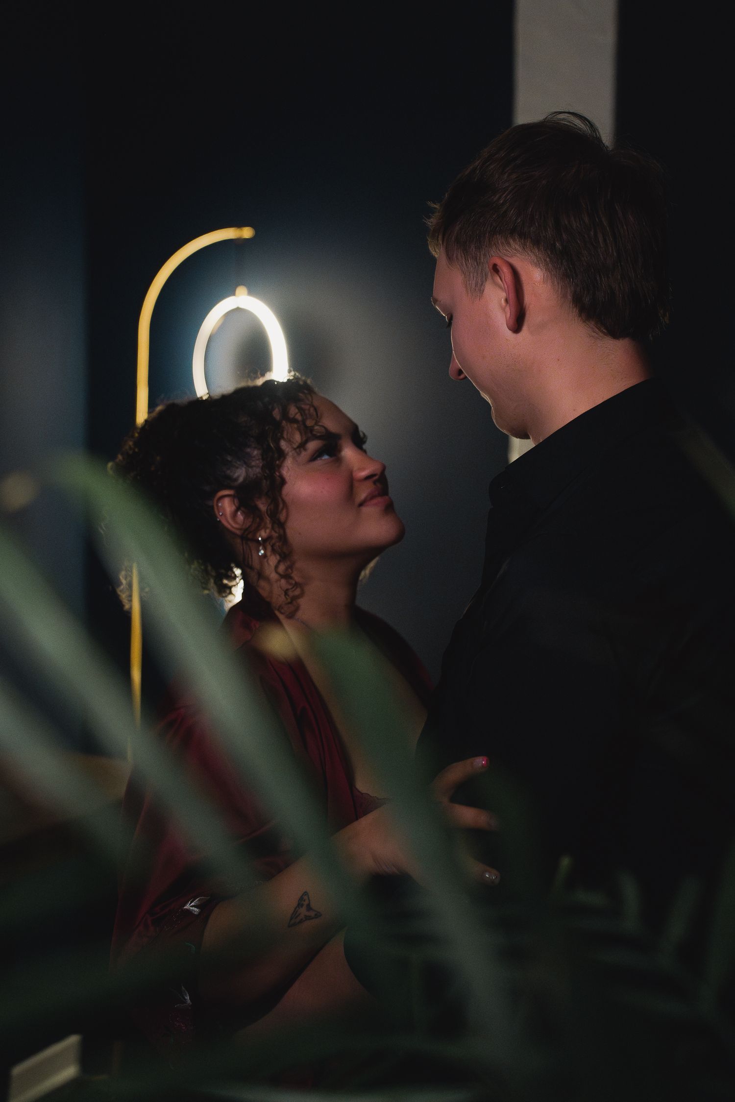 Romantic couple sharing an intimate moment in moody neon-lit bar with tropical plants in foreground.