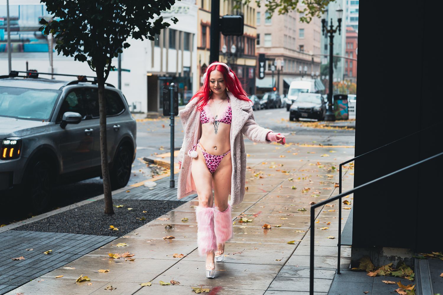 A woman with pink-red hair walks confidently in a pink leopard bikini and coat on a city sidewalk.