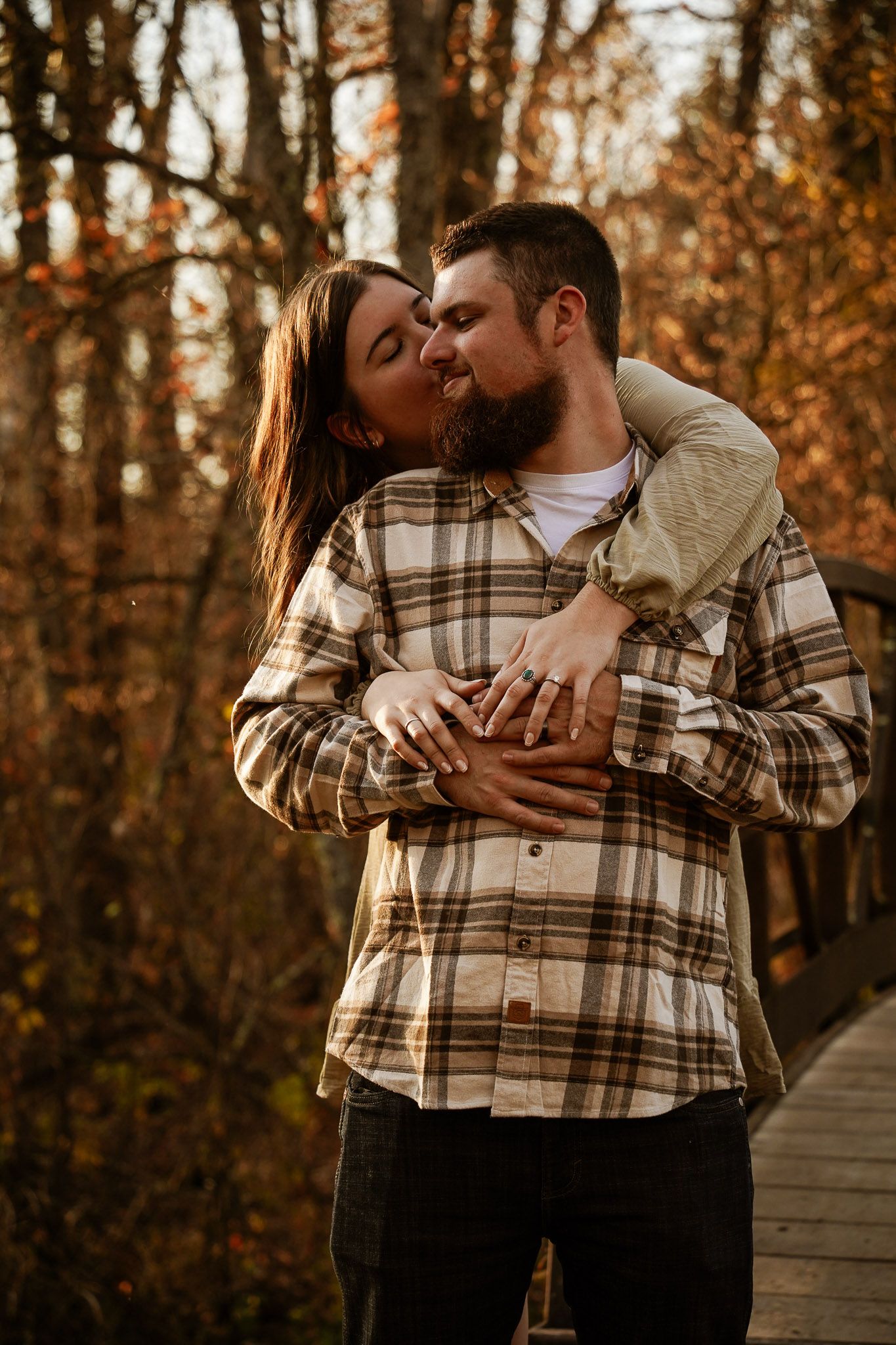 Wisconsin Couple and Engagement Photographer - Couple hugging over bridge in woods in fall during an engagement session during sunset.