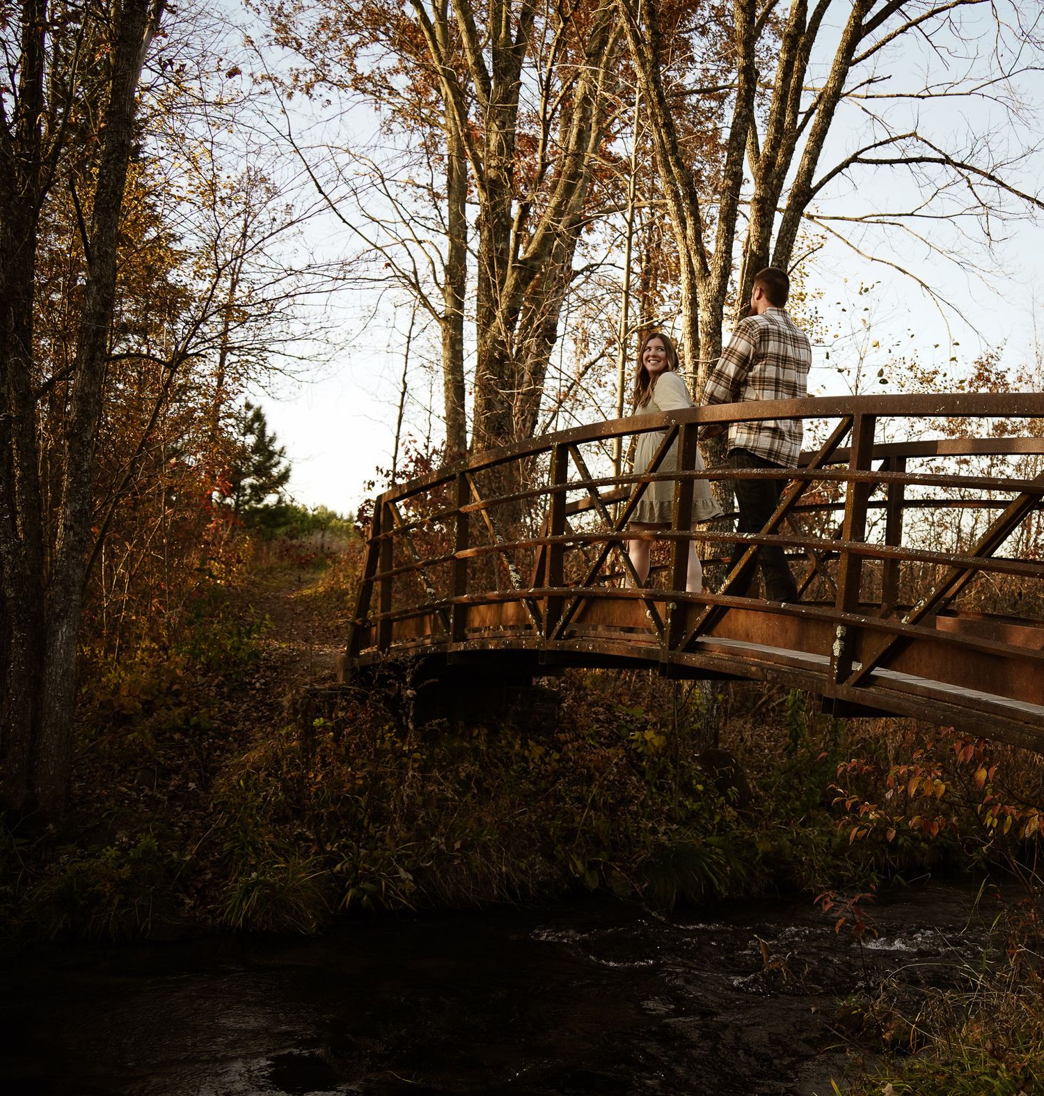 Wisconsin Couple and Engagement Photographer - Couple walking over bridge in woods in fall during an engagement session during sunset.