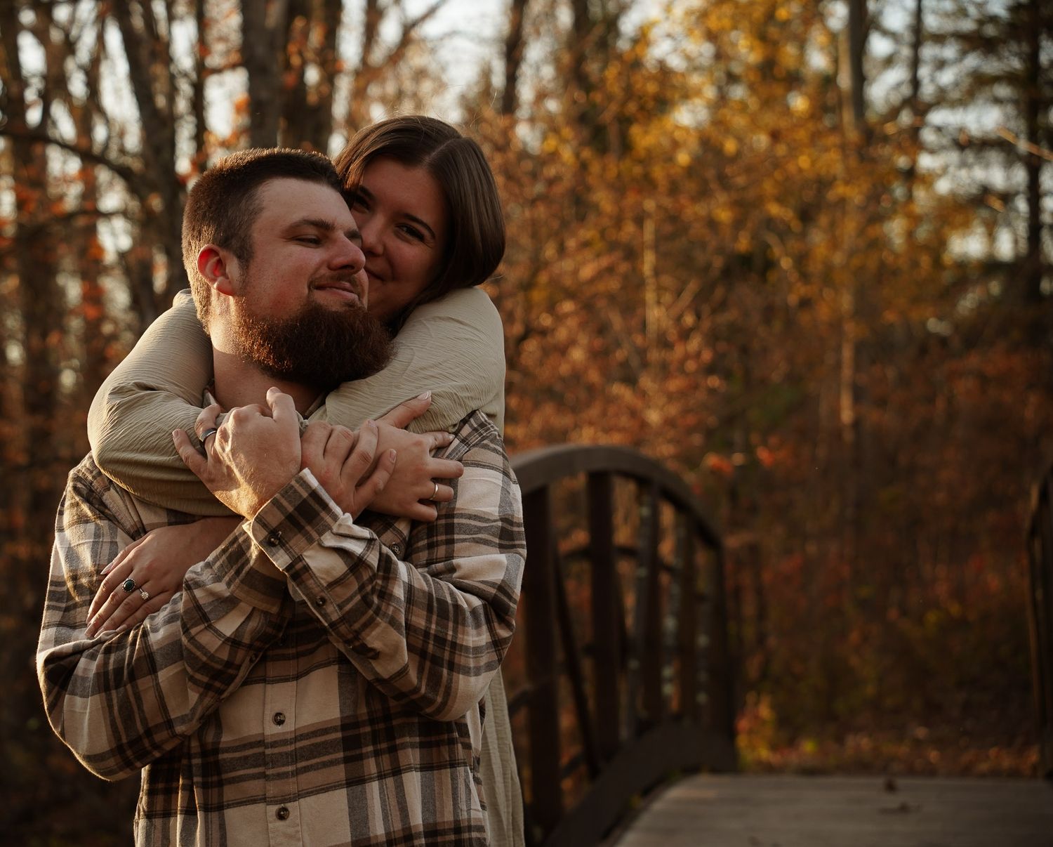 Wisconsin Couple and Engagement Photographer - Couple hugging over bridge in woods in fall during an engagement session during sunset.