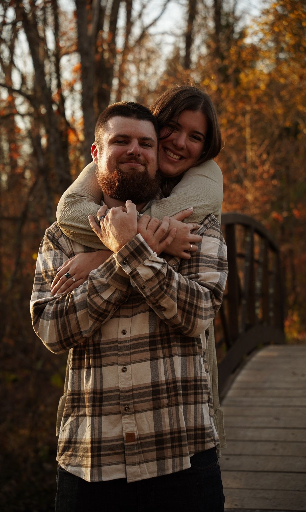 Wisconsin Couple and Engagement Photographer - Couple hugging over bridge in woods in fall during an engagement session during sunset.