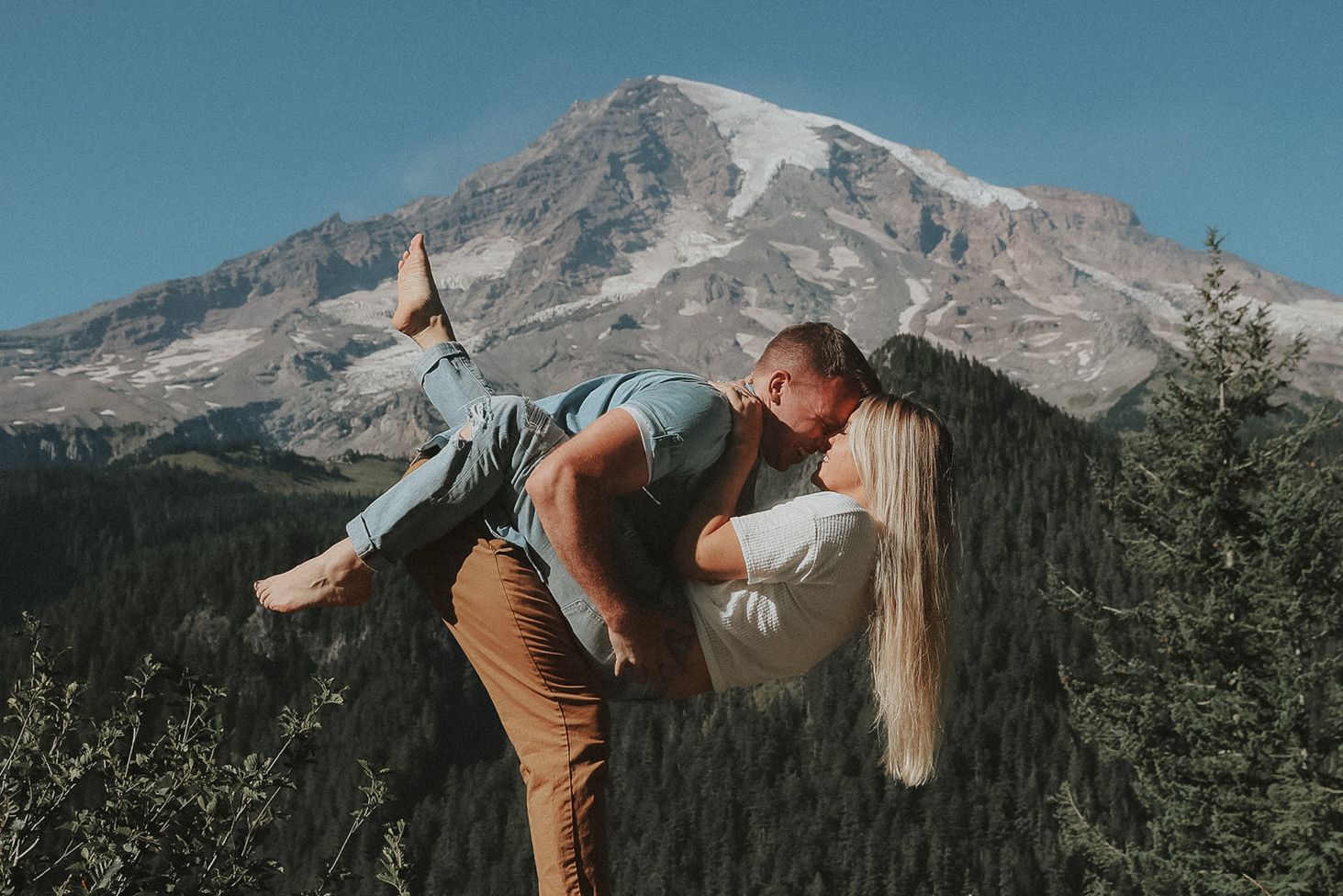 A playful engaged couple poses against a backdrop of snow-capped mountains and evergreen forest.
