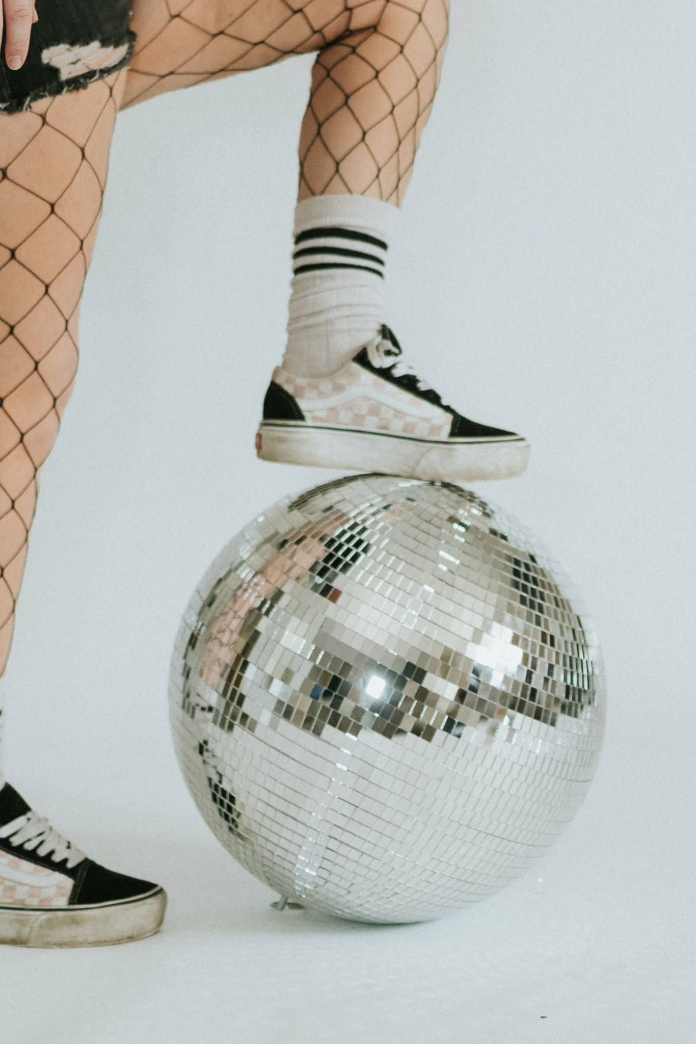 Feet wearing fishnet stockings and black sneakers stand atop a silver disco ball against a white background.
